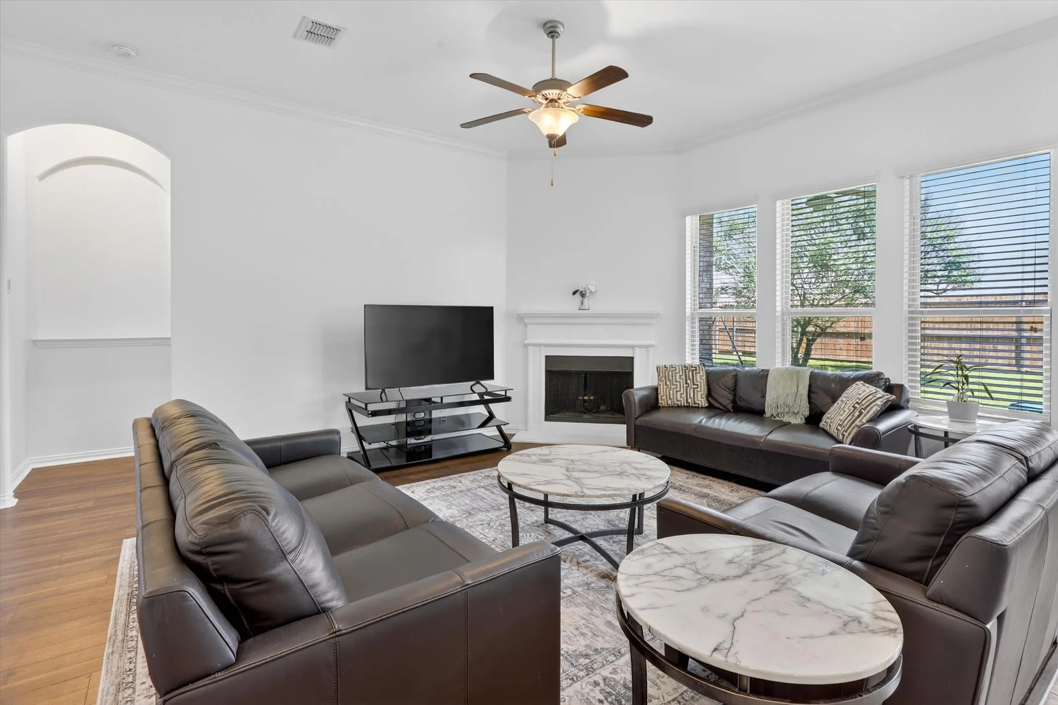 Spacious living area featuring high ceilings, wood-finish flooring, a corner fireplace with white mantle, multiple windows with horizontal blinds, and a ceiling fan with integrated lighting