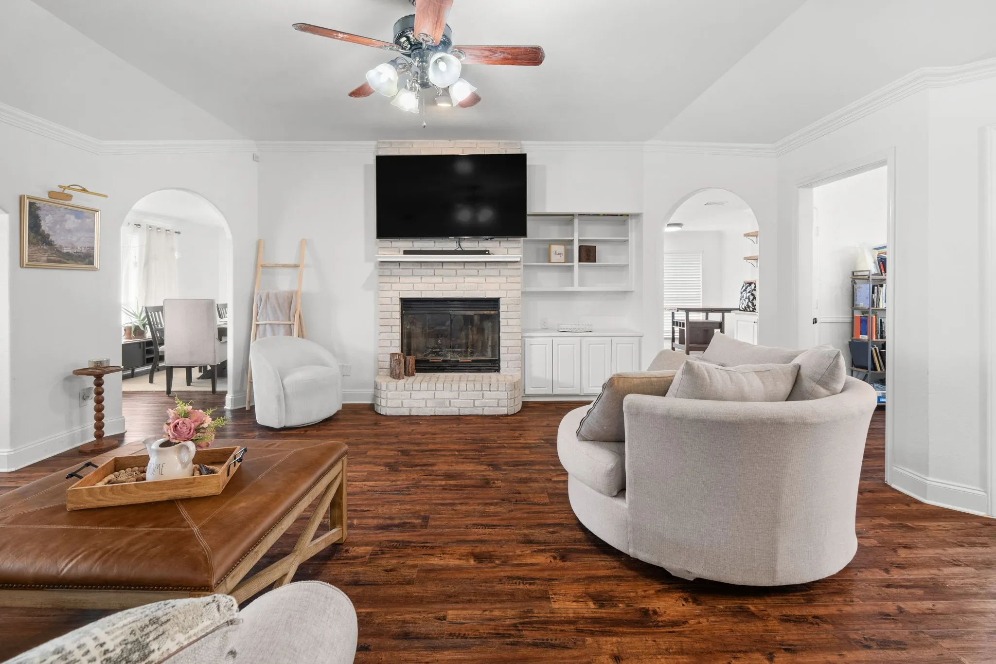 Living room featuring arched walkways, crown molding, dark wood-style floors, ceiling fan, and a brick fireplace