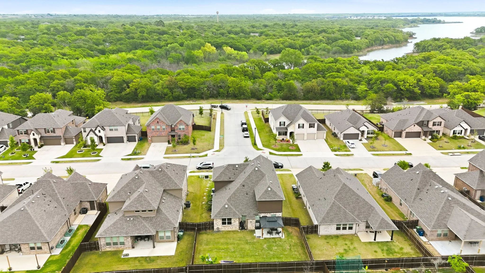Residential street with multiple homes featuring attached garages and private yards, bordered by a substantial wooded area and a body of water