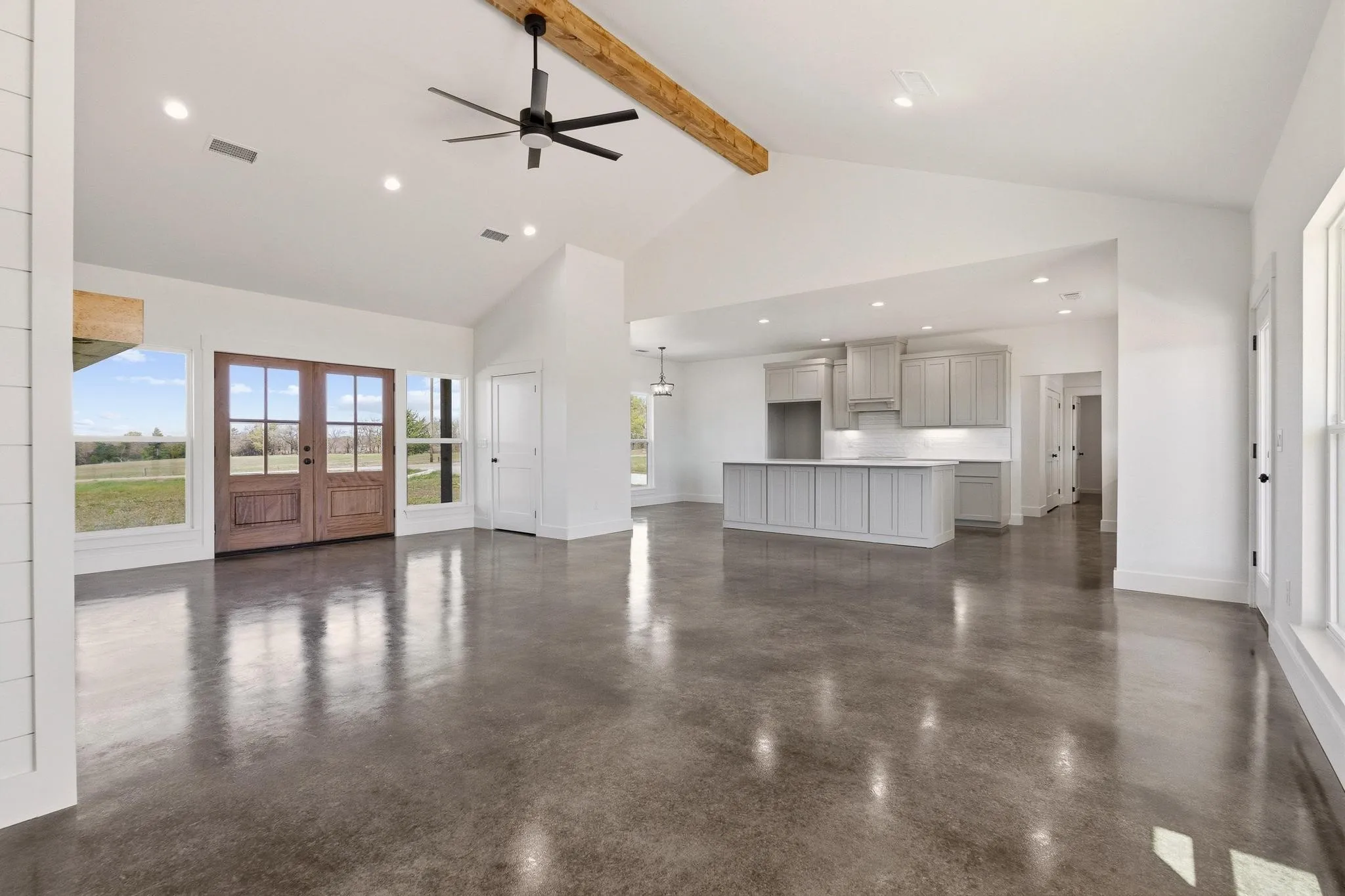 This expansive open-concept living area features polished concrete floors, a vaulted ceiling with a substantial wooden beam, and a ceiling fan
