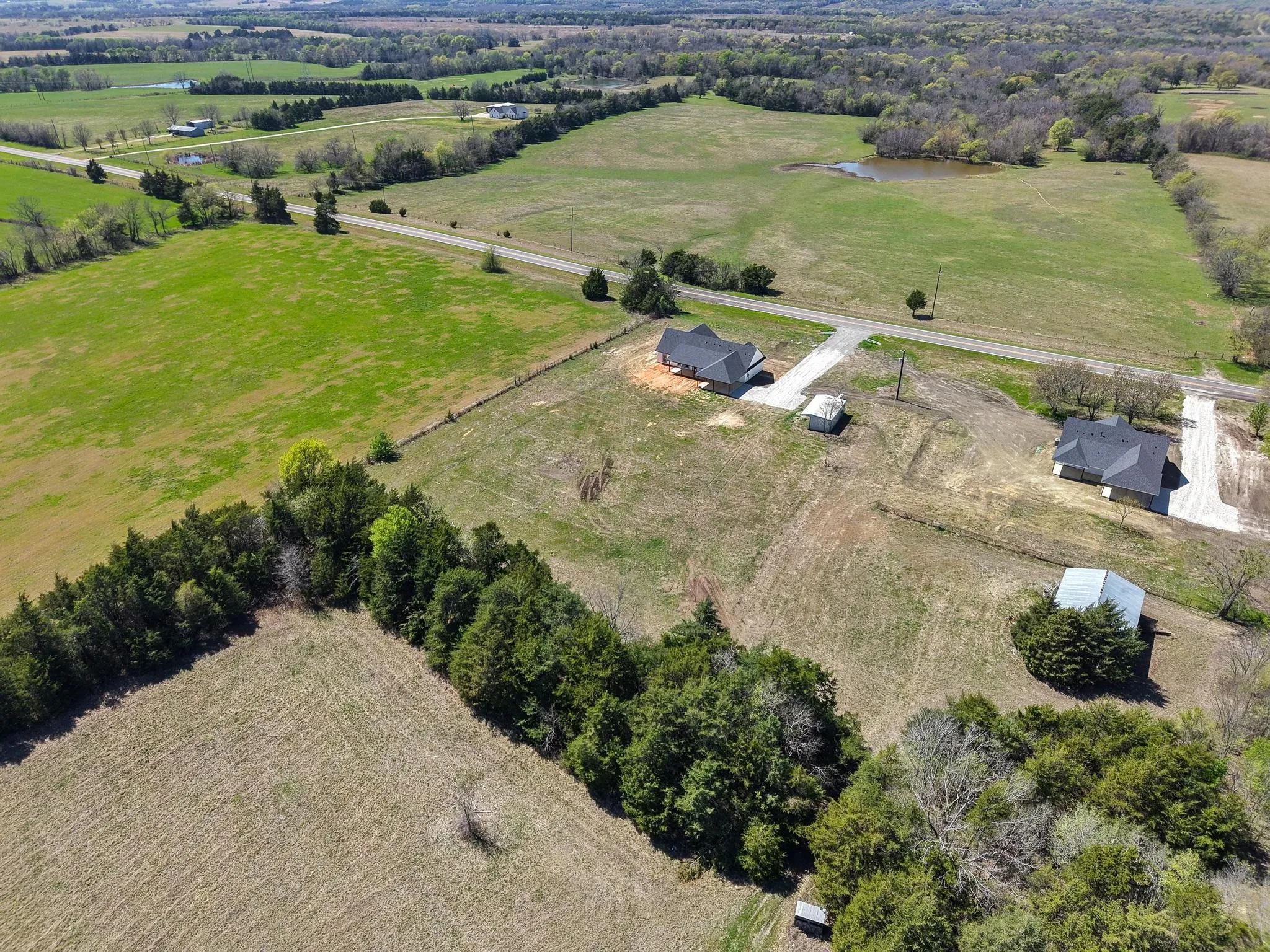 Expansive property offering a blend of open fields and wooded areas, featuring a large, dark-roofed structure, and a smaller, light-roofed outbuilding