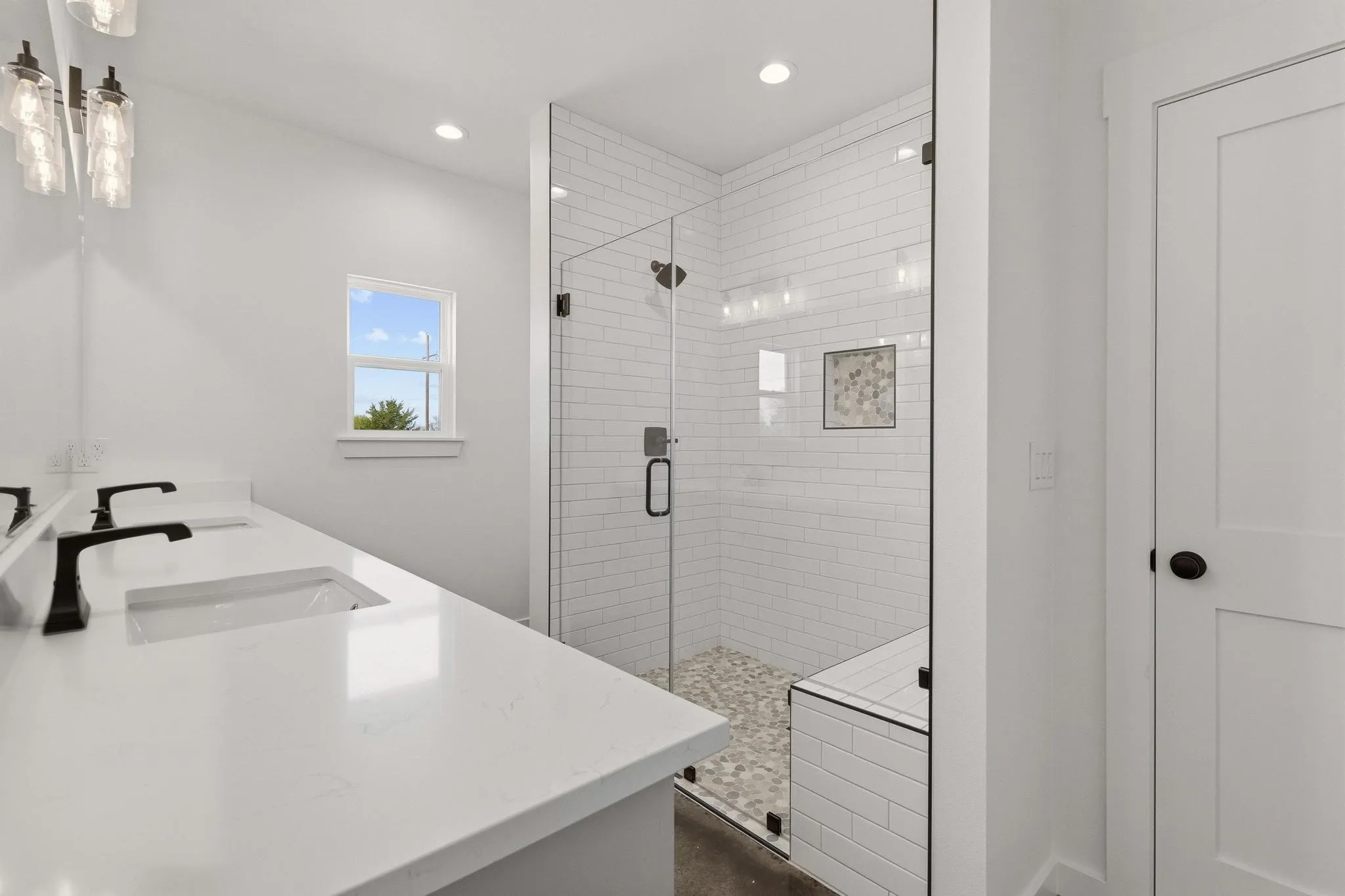 Bathroom featuring a double vanity with matte black faucets, a tiled shower with a glass enclosure, a built-in shower bench, and a window providing natural light