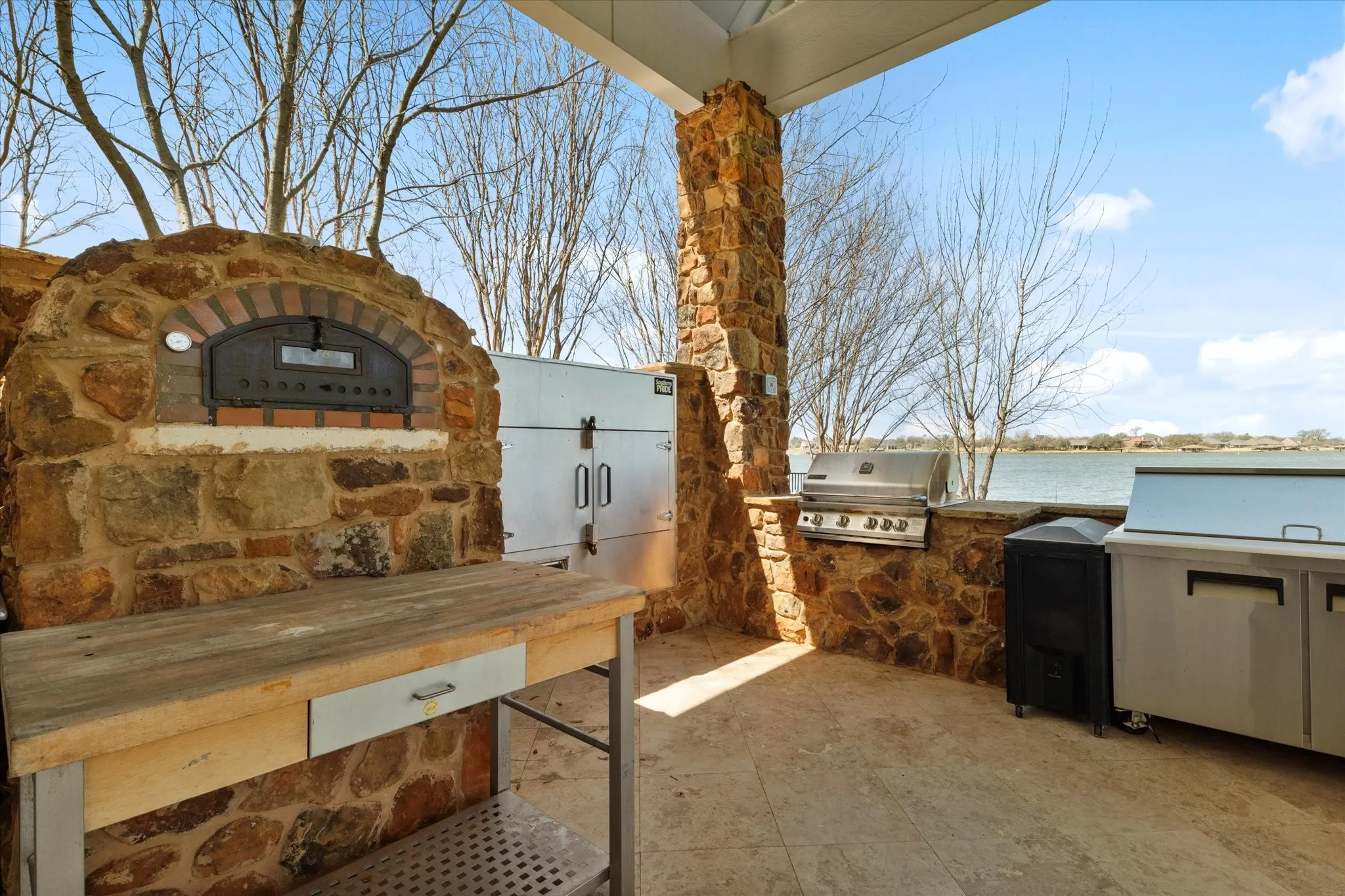 Outdoor kitchen featuring a stone-clad pizza oven, a built-in grill, and stainless steel appliances, all set on a tiled patio