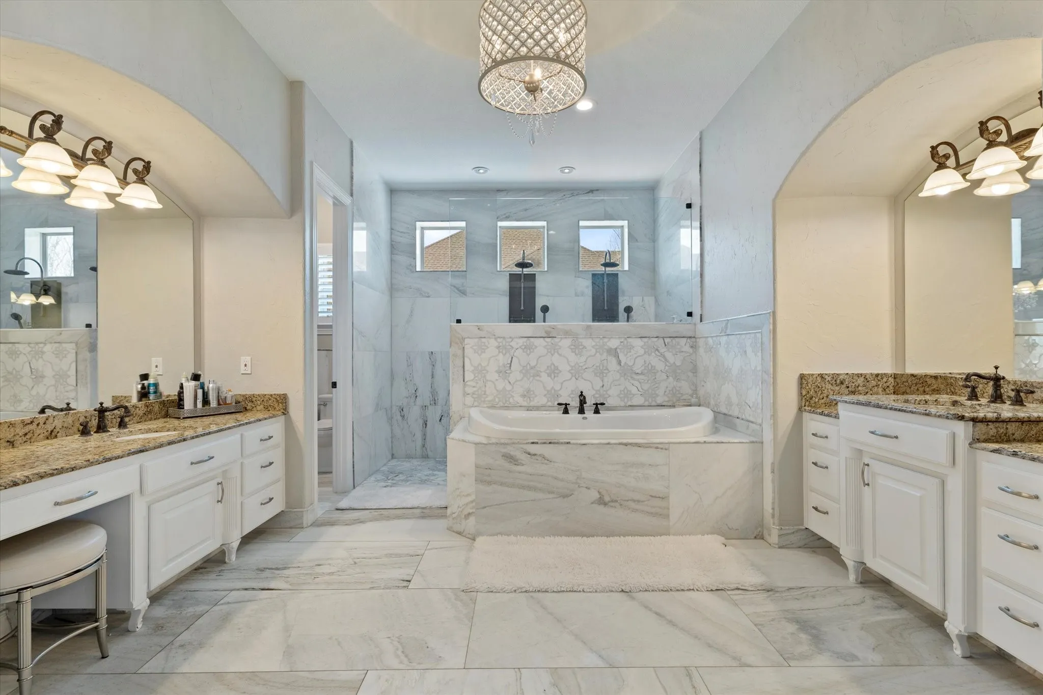 Luxurious bathroom featuring extensive marble-look tile, a large soaking tub, and dual vanity areas with granite countertops