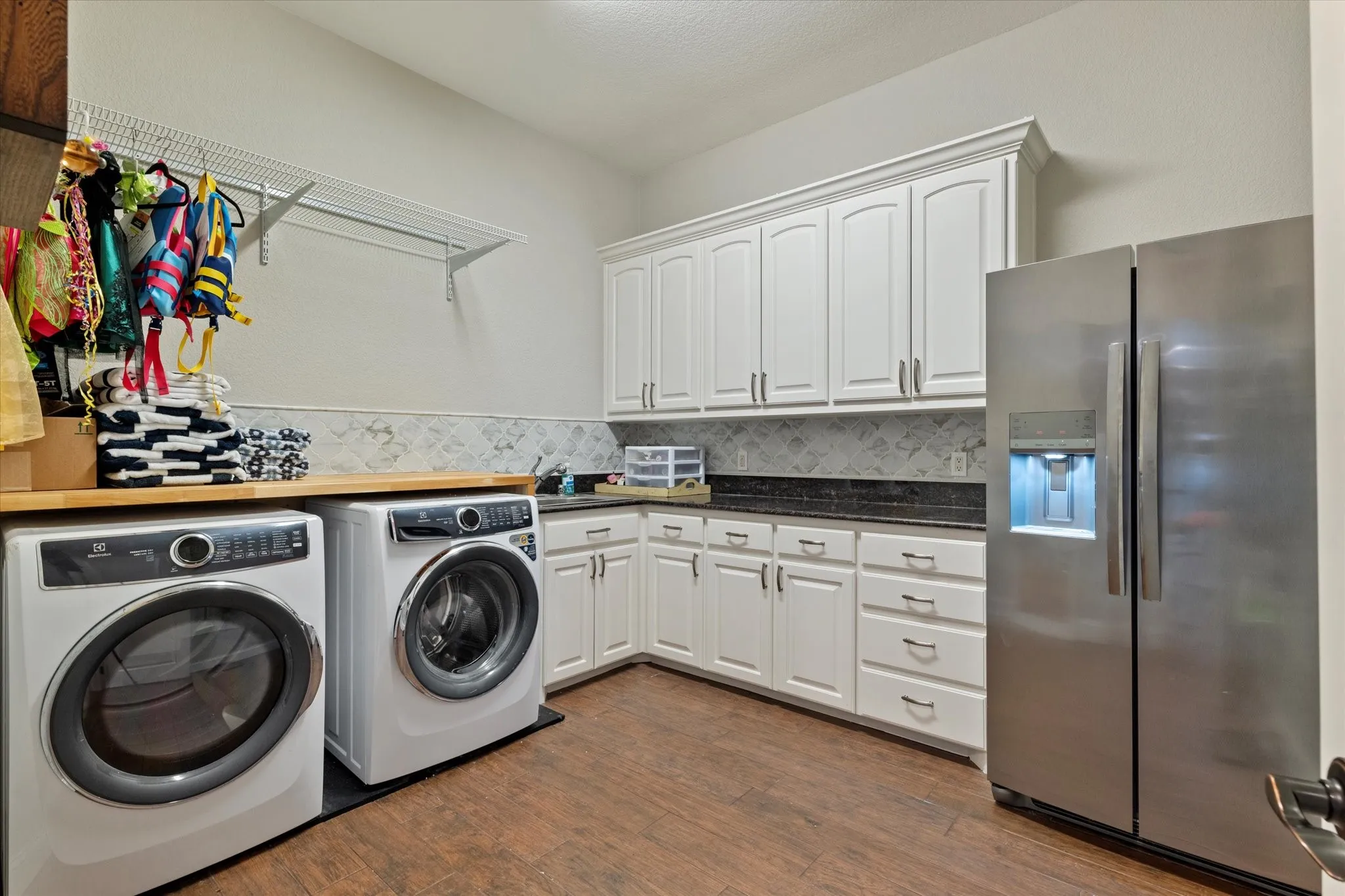 This utility area features white cabinetry with dark countertops, a stainless steel refrigerator, and wood-look flooring