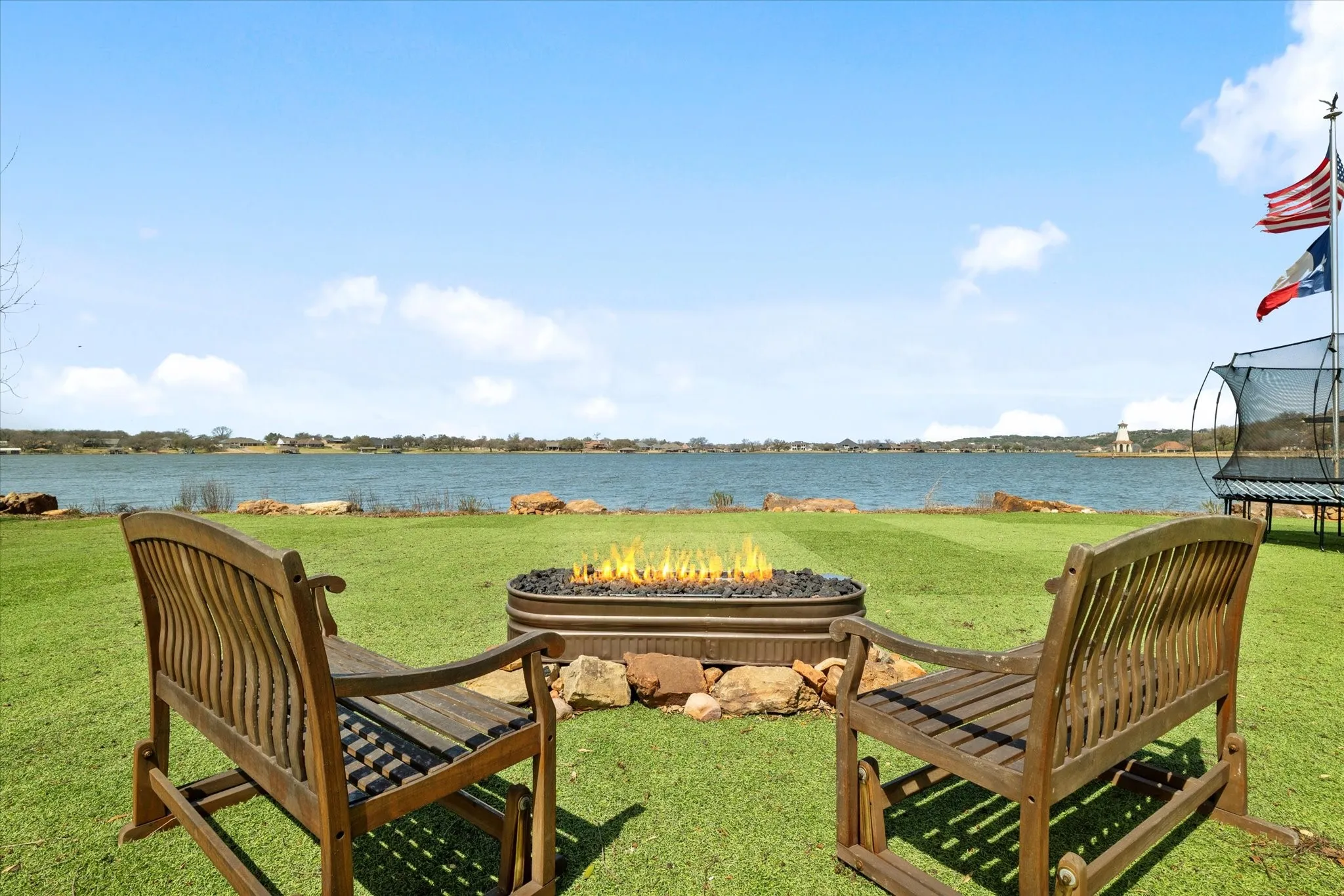 The property features a spacious lawn with a fire pit, two wooden benches, and a large body of water in the background