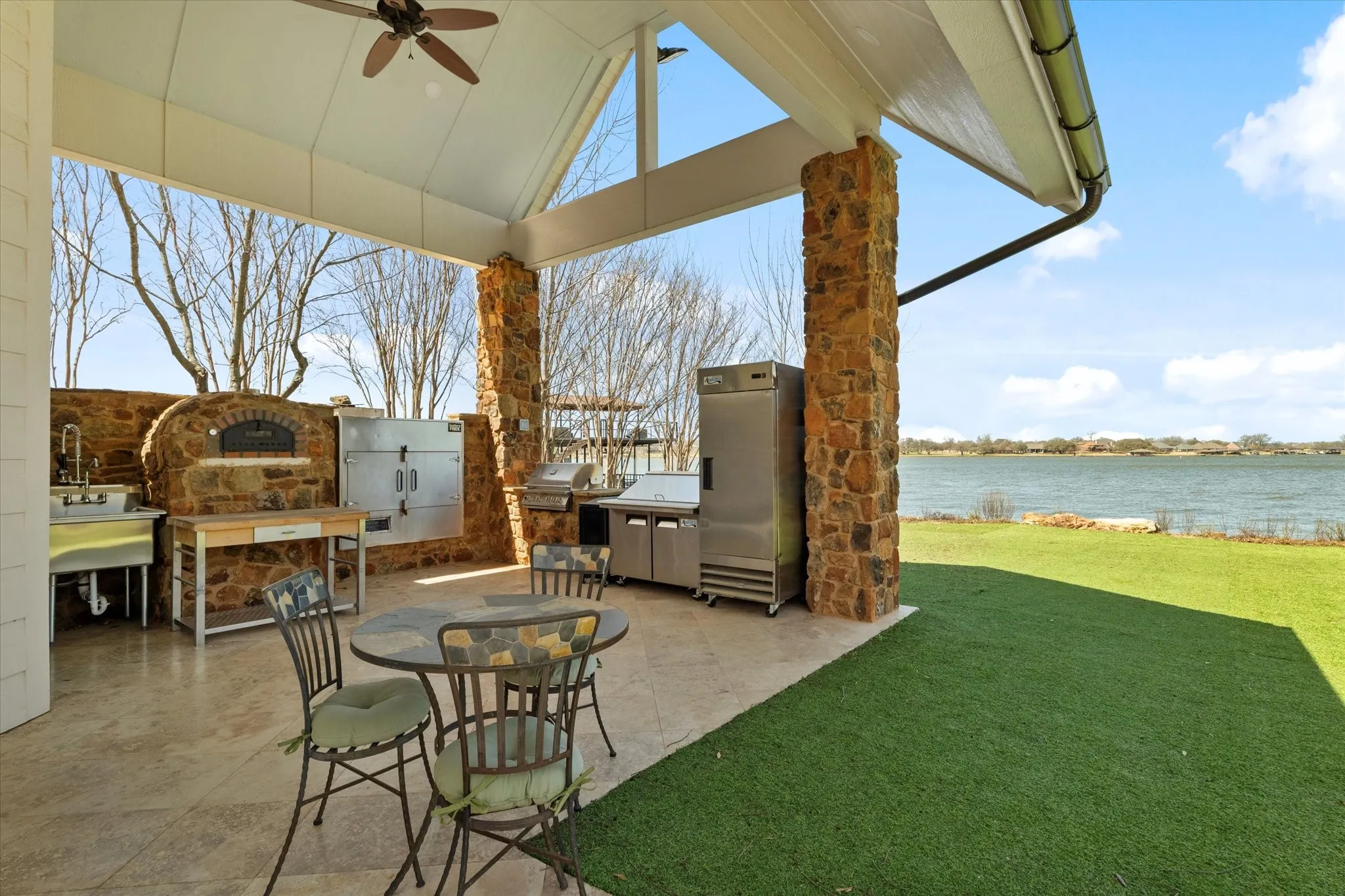 Outdoor living area featuring a covered patio with stone columns, an outdoor kitchen with a built-in pizza oven, and a green lawn overlooking a body of water