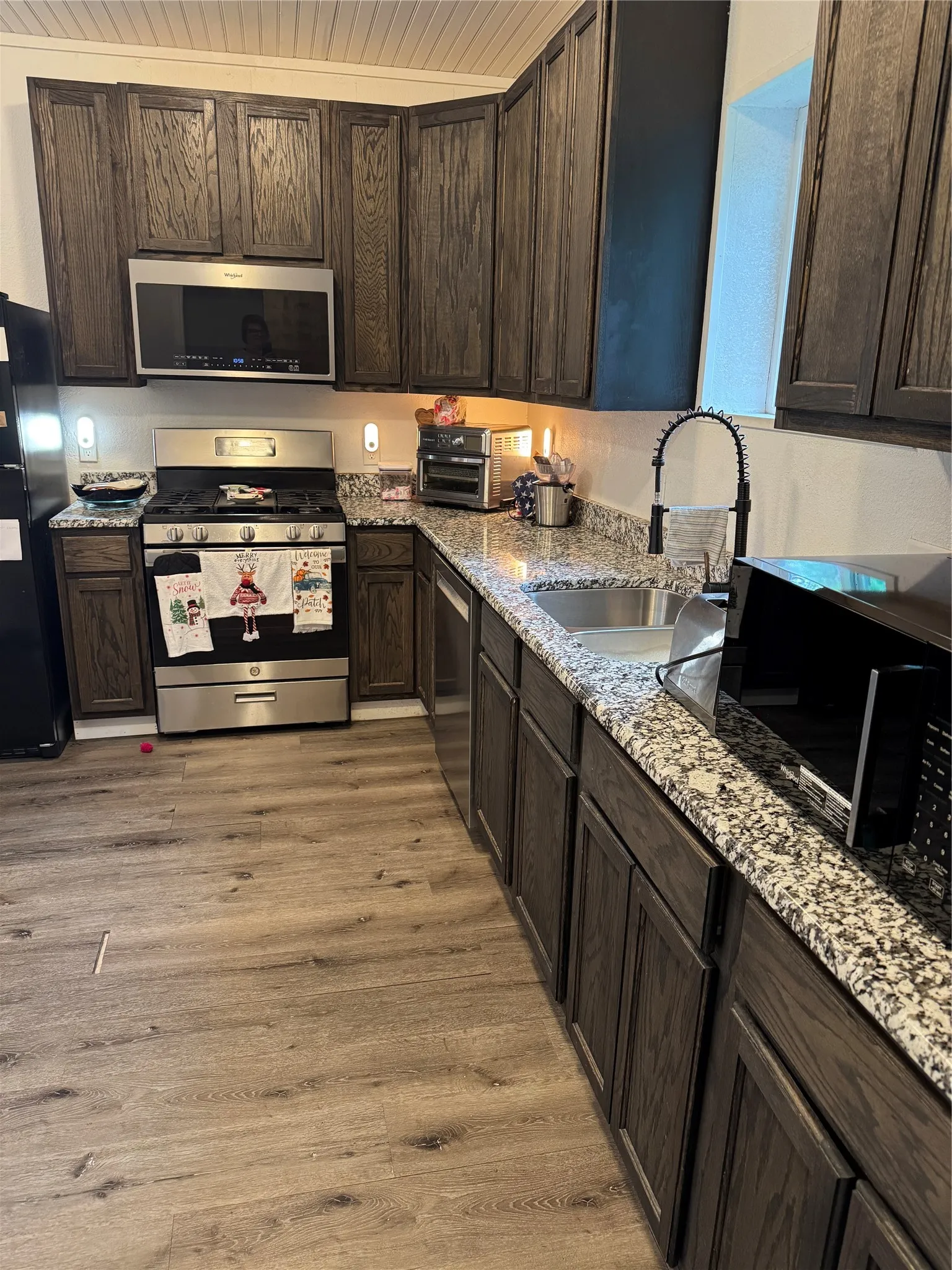 Kitchen featuring dark wood finish cabinets, black appliances, light stone countertops, light wood-type flooring, and wooden ceiling
