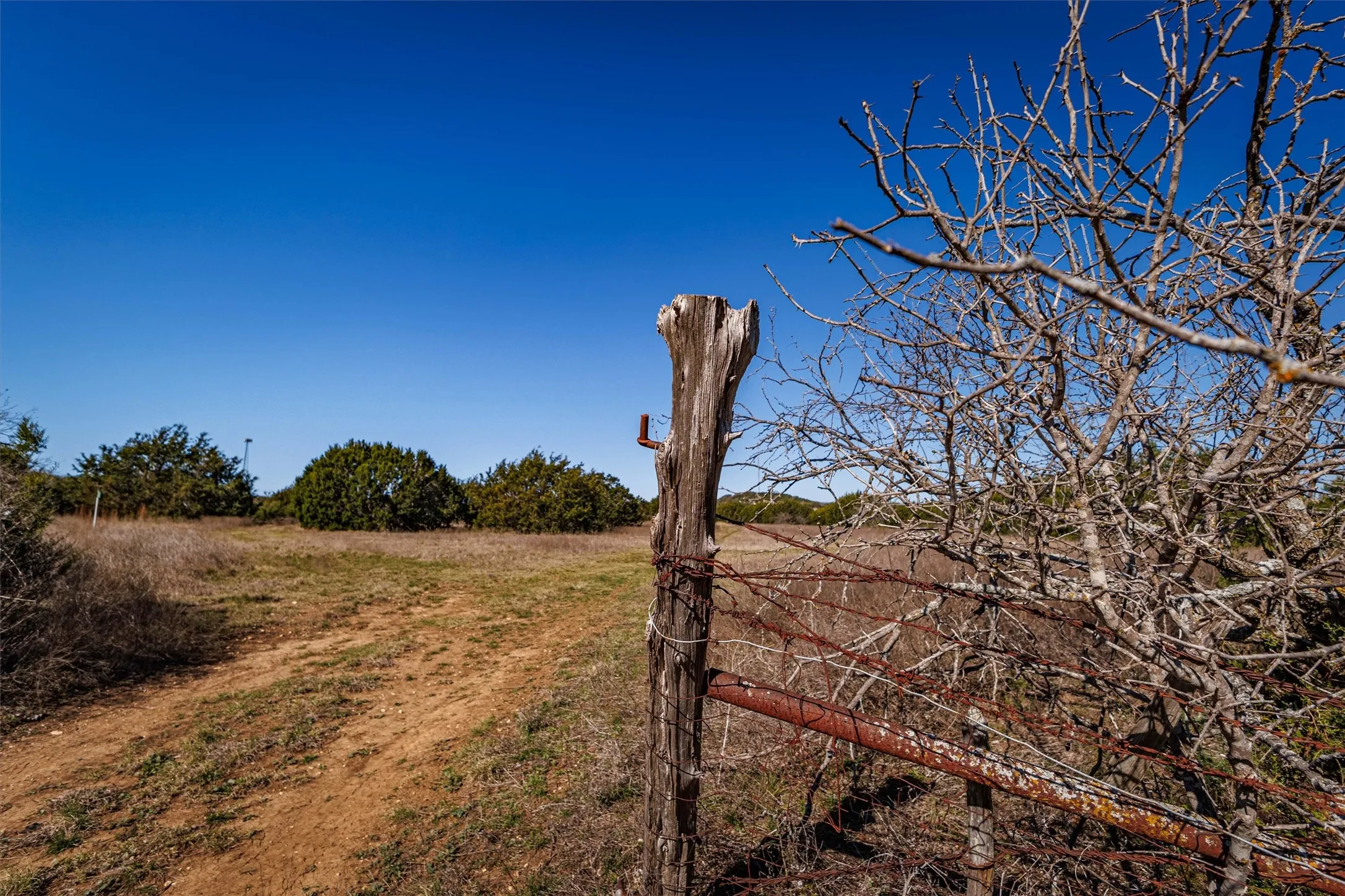 The property features a clear blue sky, a rural landscape with a dirt path, and a wooden fence post with barbed wire