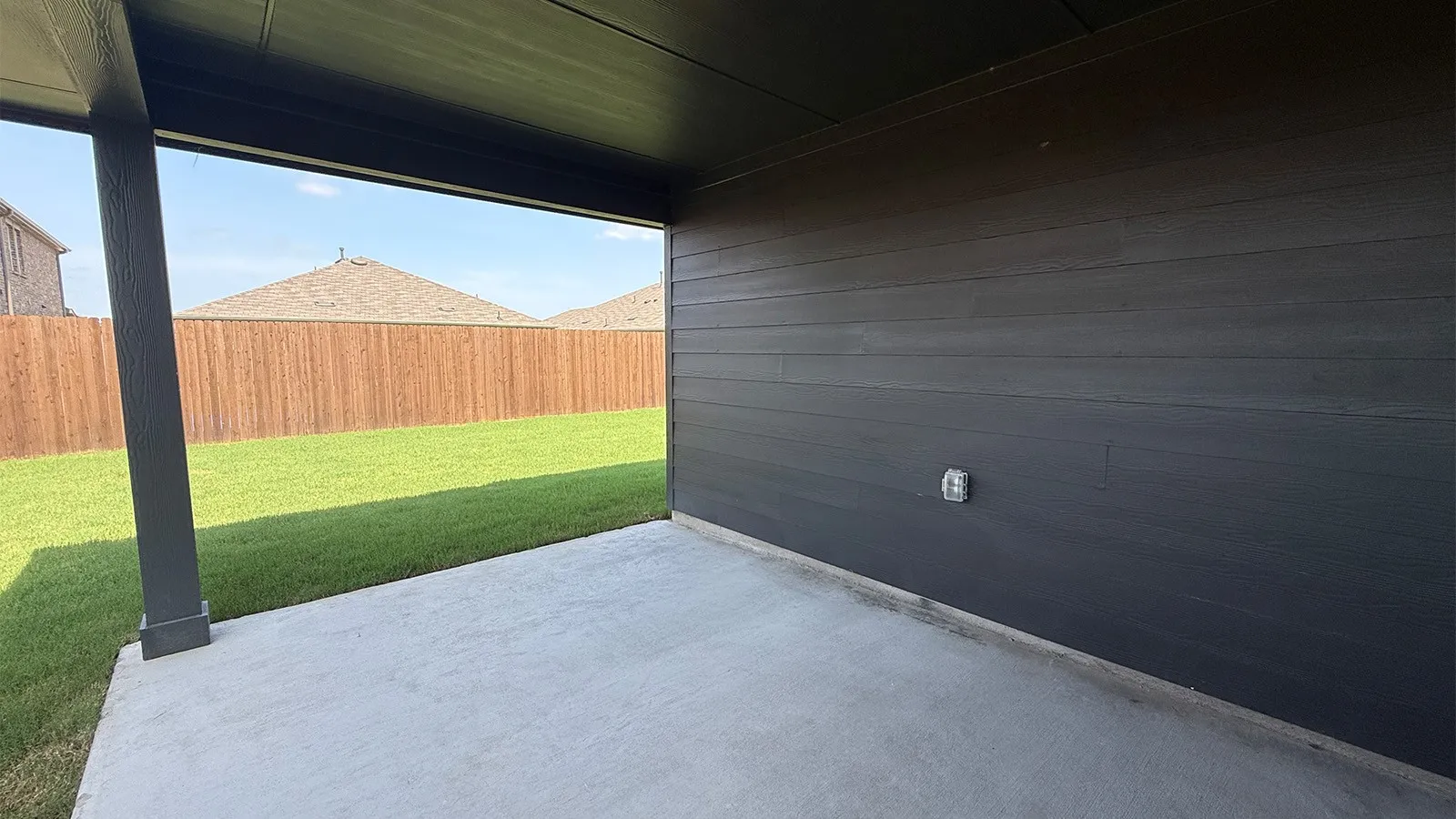 Covered concrete patio with a dark wood-finish ceiling, dark horizontal siding, and an electrical outlet
