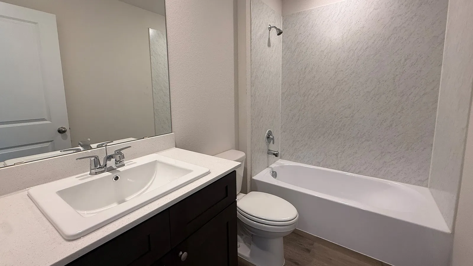 Bathroom featuring a dark wood-finish vanity with a white countertop and integrated sink, a large wall-mounted mirror, a tub/shower combination with a textured surround, and wood-finish flooring