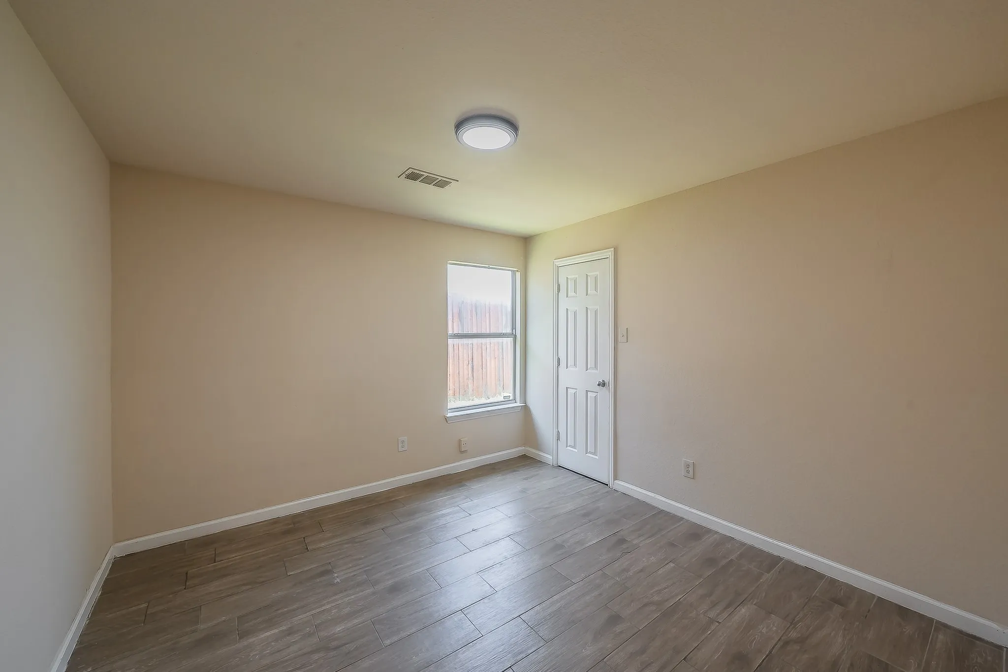 Spacious room featuring wood-finish flooring, neutral-toned walls, a single window, a white paneled door, and a flush-mount ceiling light fixture