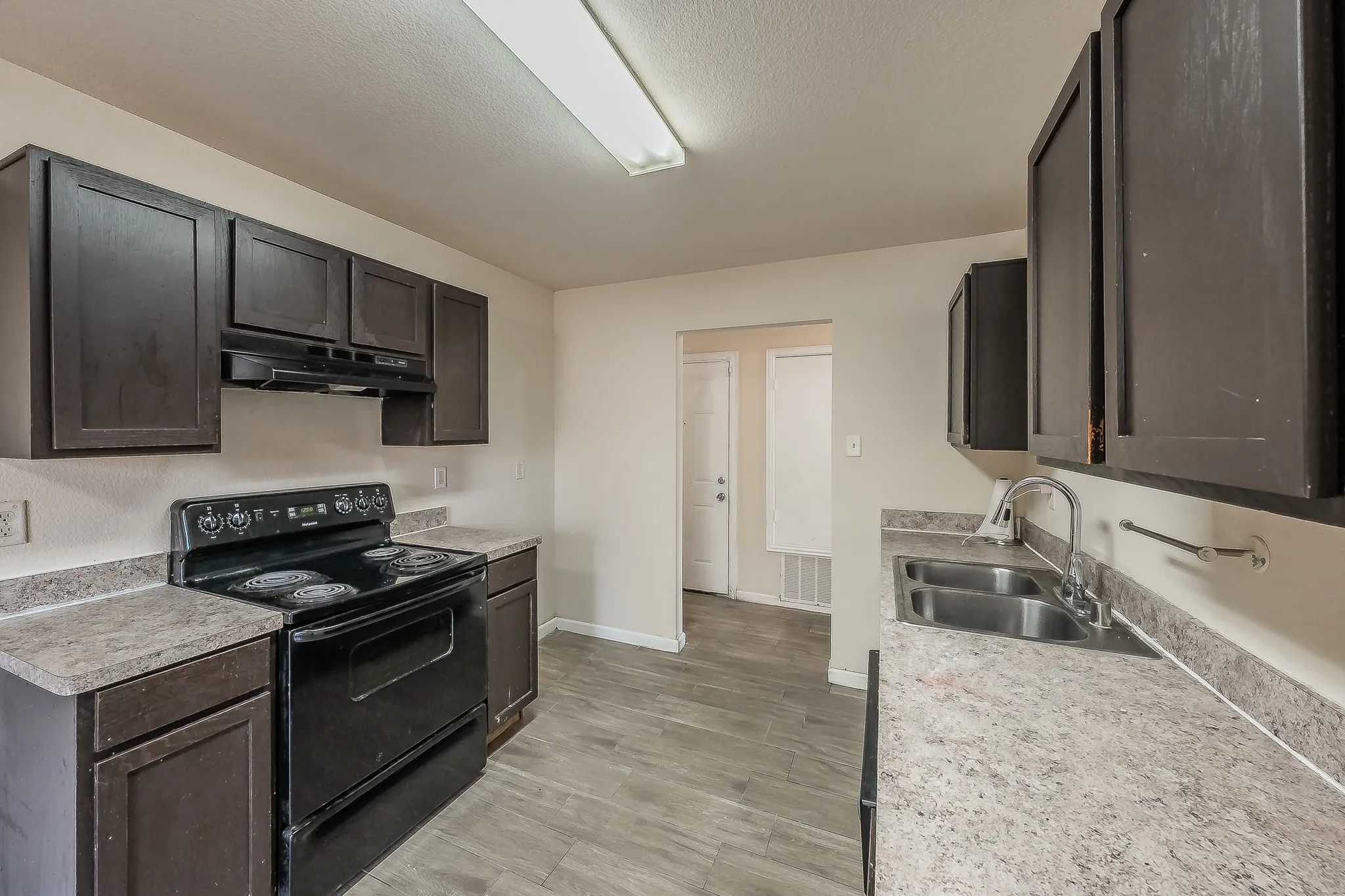 Kitchen featuring dark wood-finish cabinetry, black electric range with coordinating hood, double-basin stainless steel sink with gooseneck faucet, light-toned countertop, and wood-finish flooring
