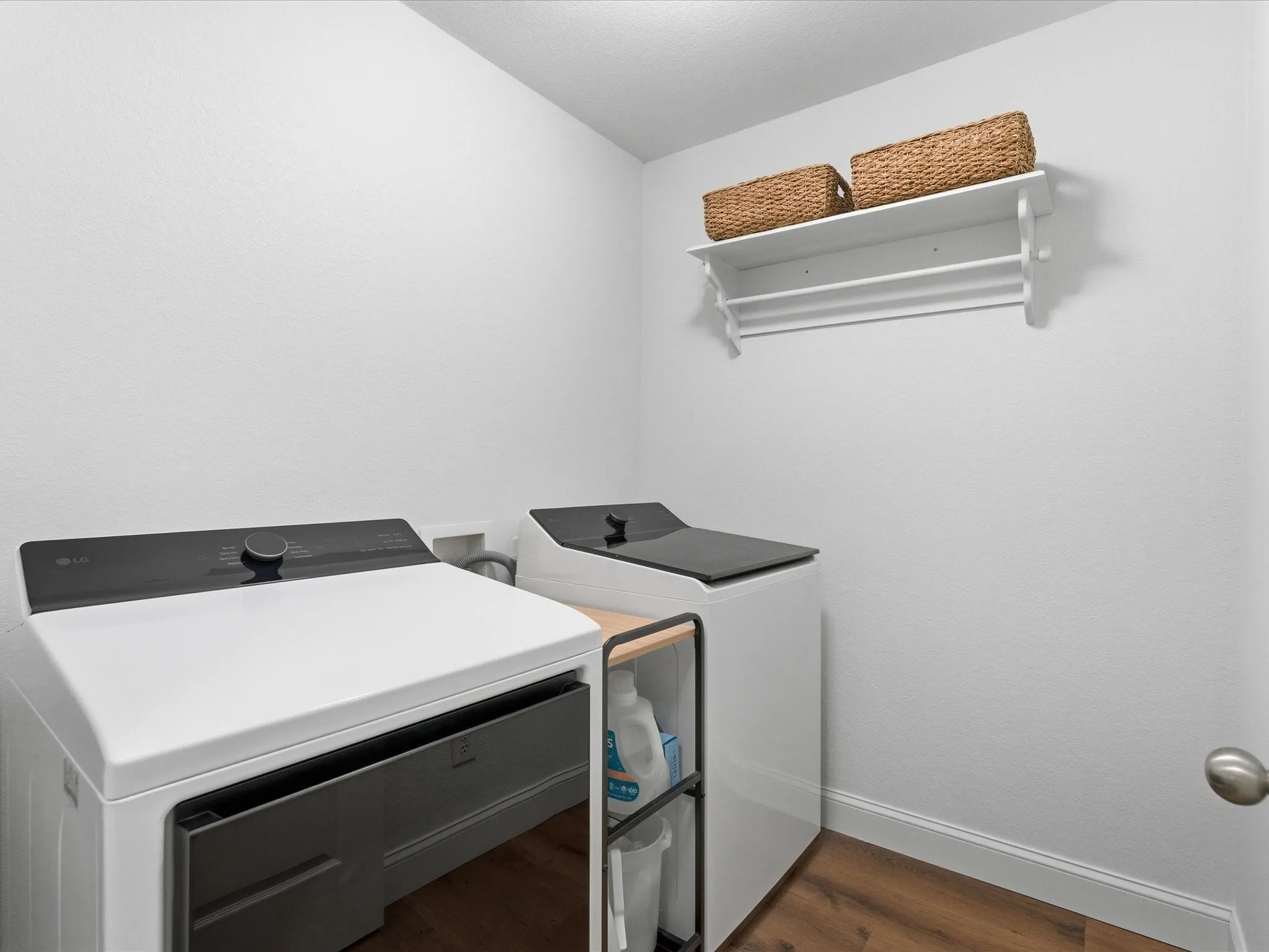 This laundry room features white walls, dark wood-style flooring, and a white wall-mounted shelf with hanging rods