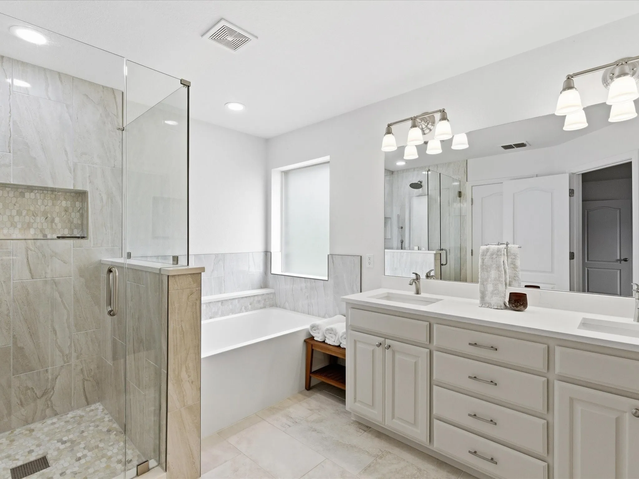 This well-appointed bathroom features a spacious glass-enclosed shower with a tiled bench and an integrated niche, a separate bathtub, and a dual vanity with ample storage