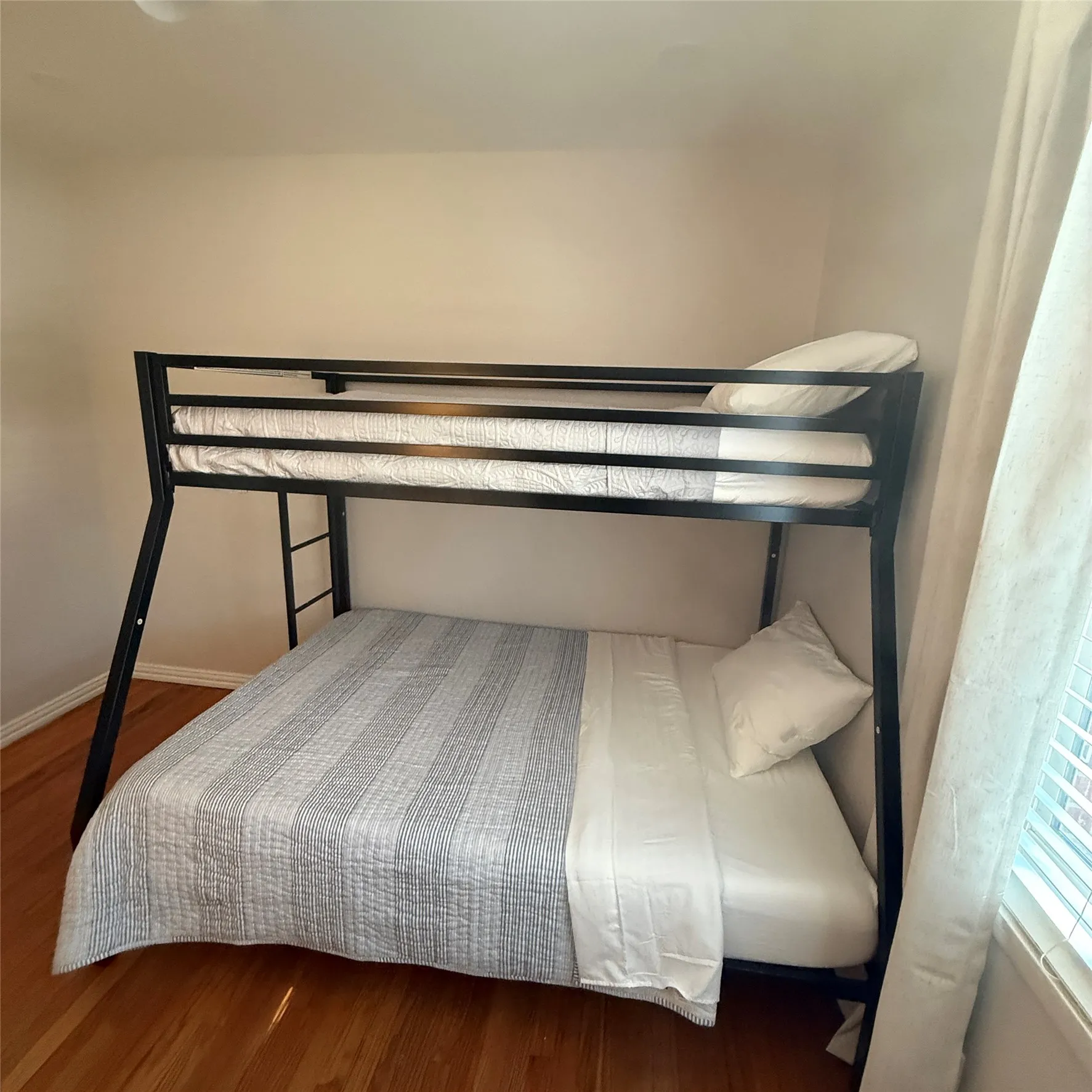 This room features warm-toned hardwood flooring, light-colored walls, and a window with blinds and curtains