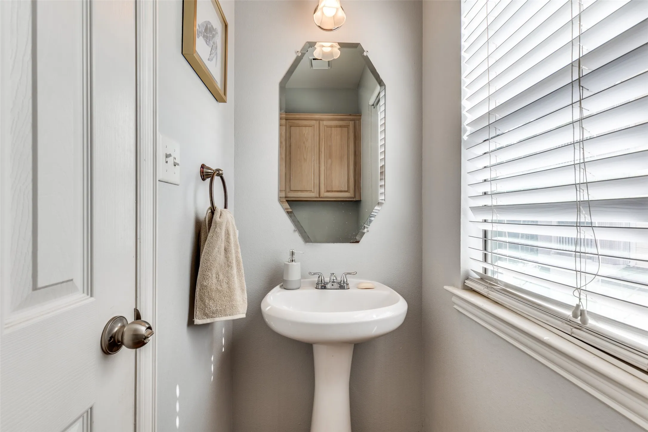 This bathroom features a white pedestal sink with a chrome faucet, an octagonal mirror, and a window with white blinds
