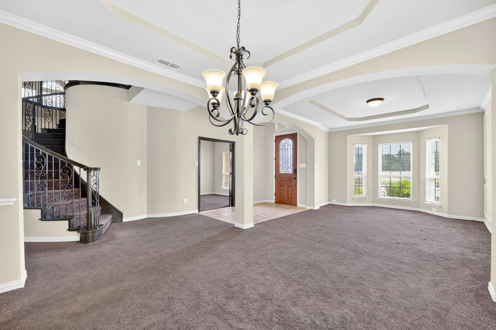 Spacious living area featuring neutral-toned carpet, a chandelier, and a curved staircase with dark iron railings