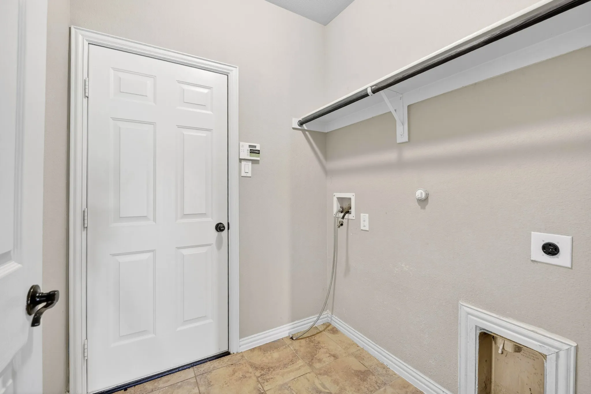This utility area features tiled flooring, a white paneled door, and a wall-mounted shelf with a hanging rod