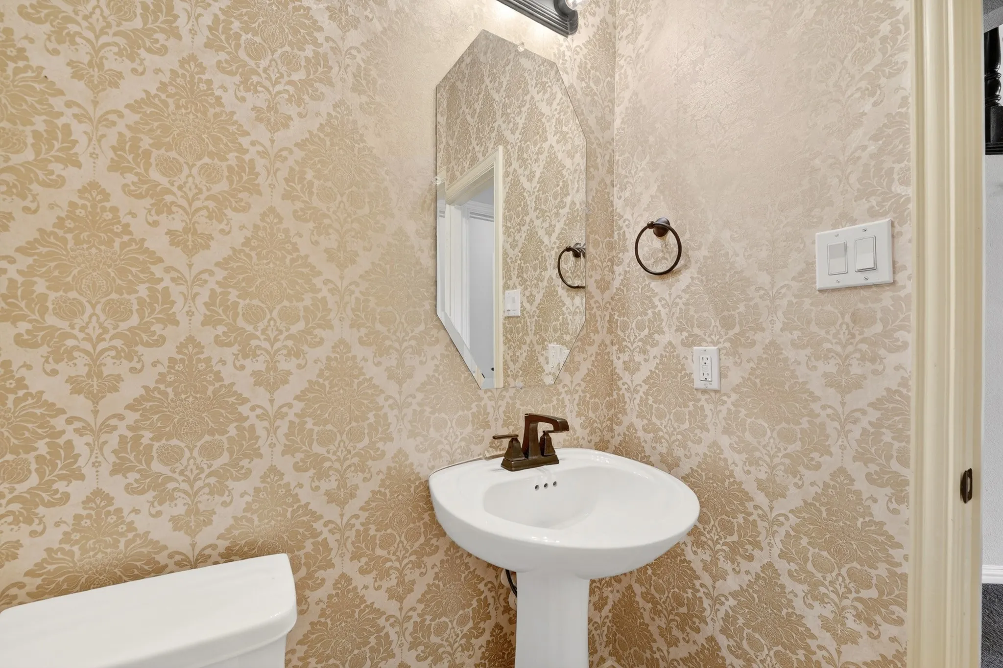This room features walls adorned with decorative wallpaper, a white pedestal sink with a bronze faucet, and a toilet