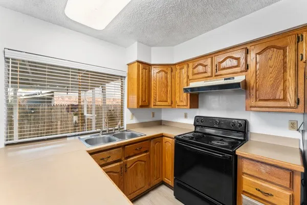 Kitchen with electric range, brown cabinetry, light countertops, a textured ceiling, and under cabinet range hood