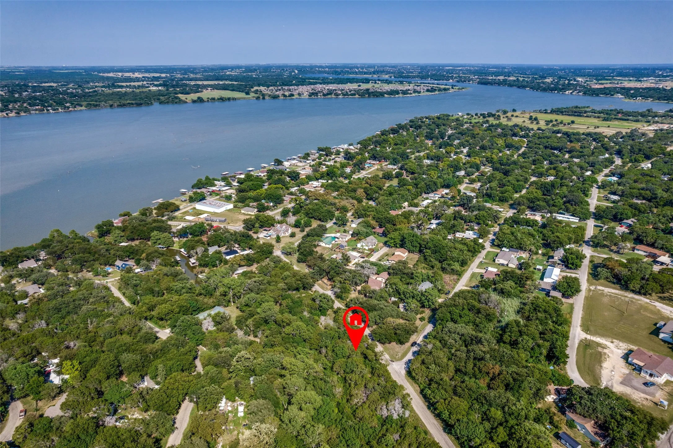 Aerial view of the property with a visible red location marker, surrounded by lush green trees and a body of water in the background
