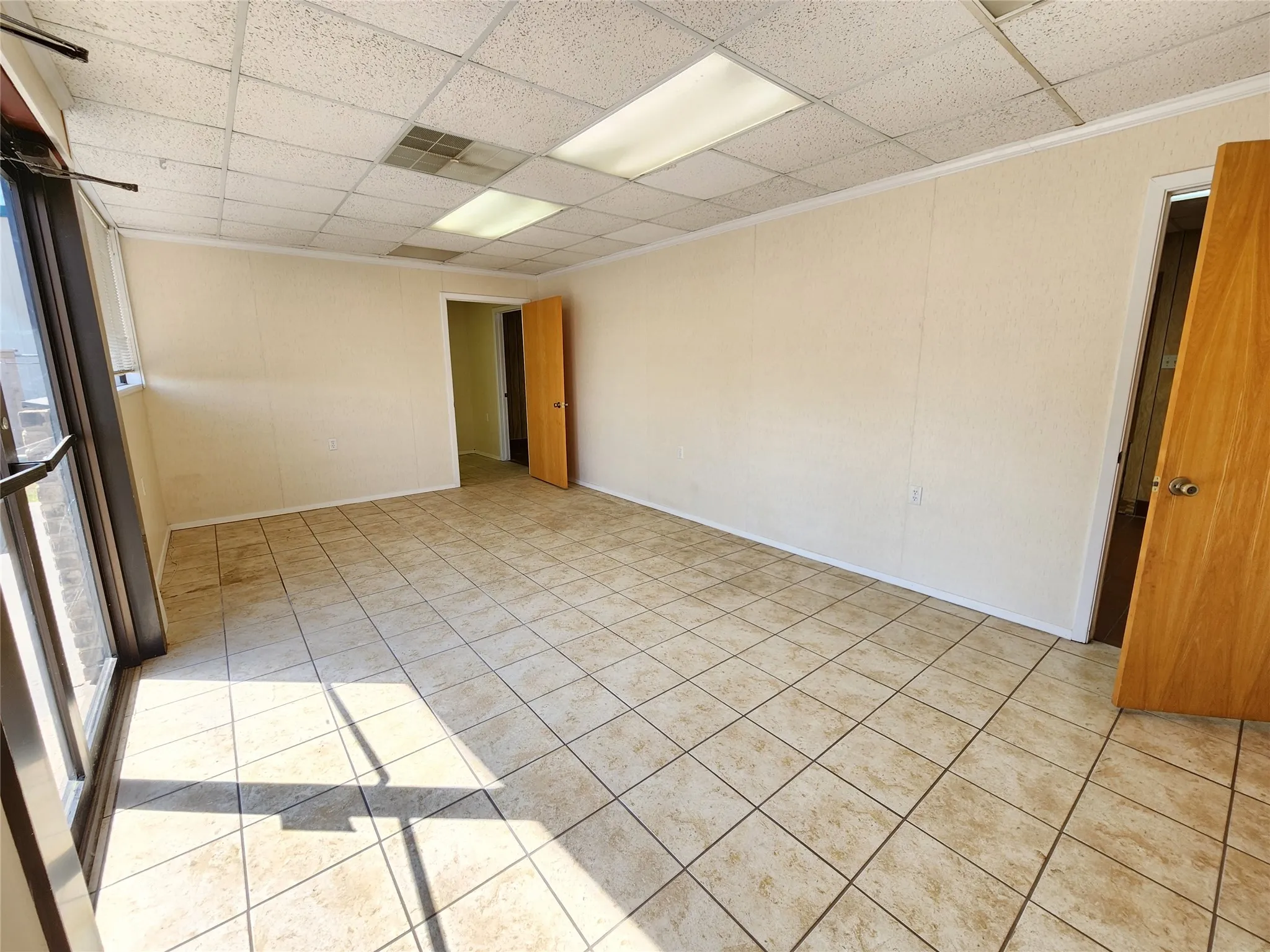 Spacious room featuring light-colored tile flooring, neutral-toned walls, and a drop ceiling with integrated lighting