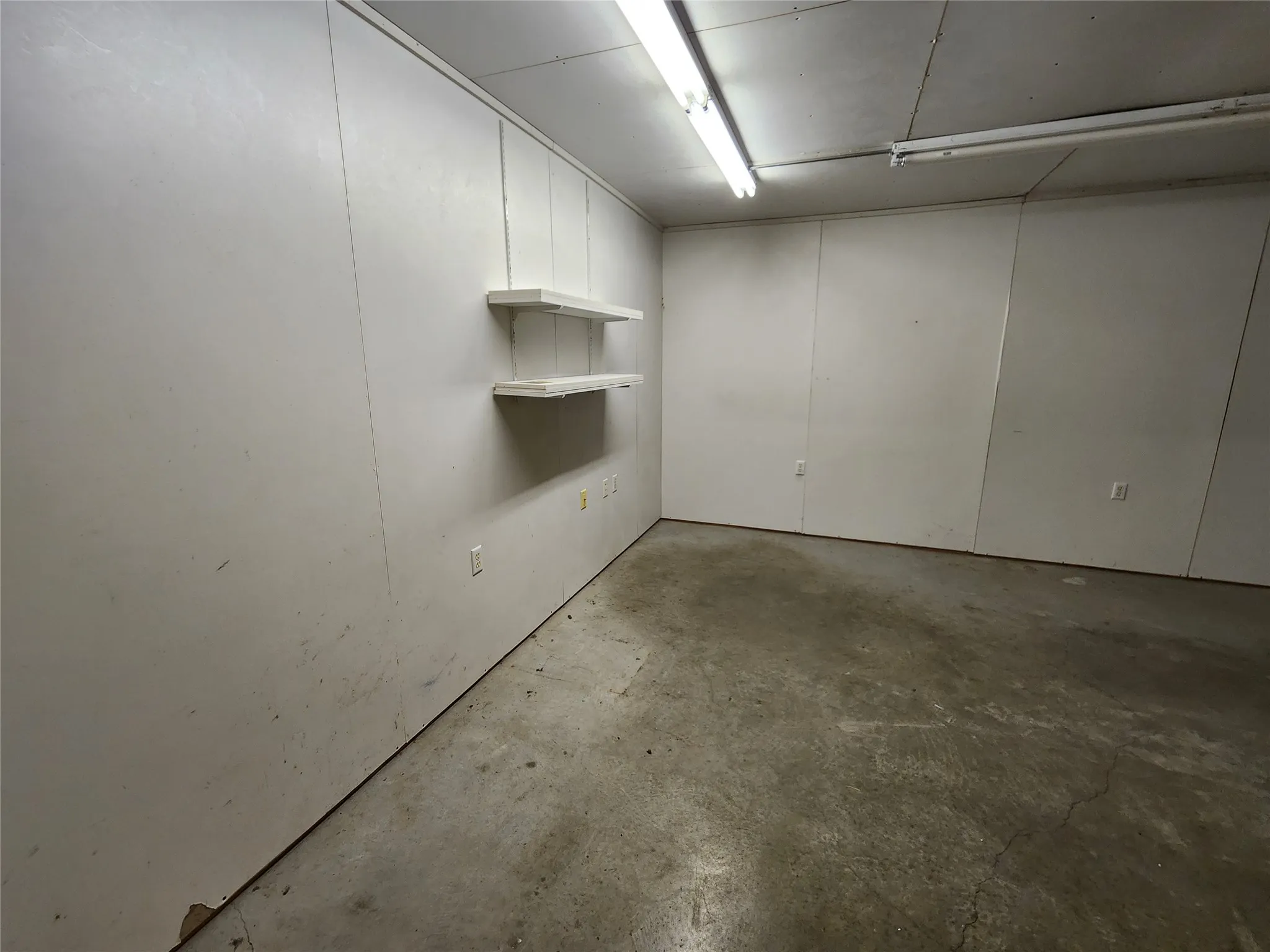 The property features a utility room with white paneling walls, two white wall-mounted shelves, a concrete floor, and fluorescent lighting fixtures