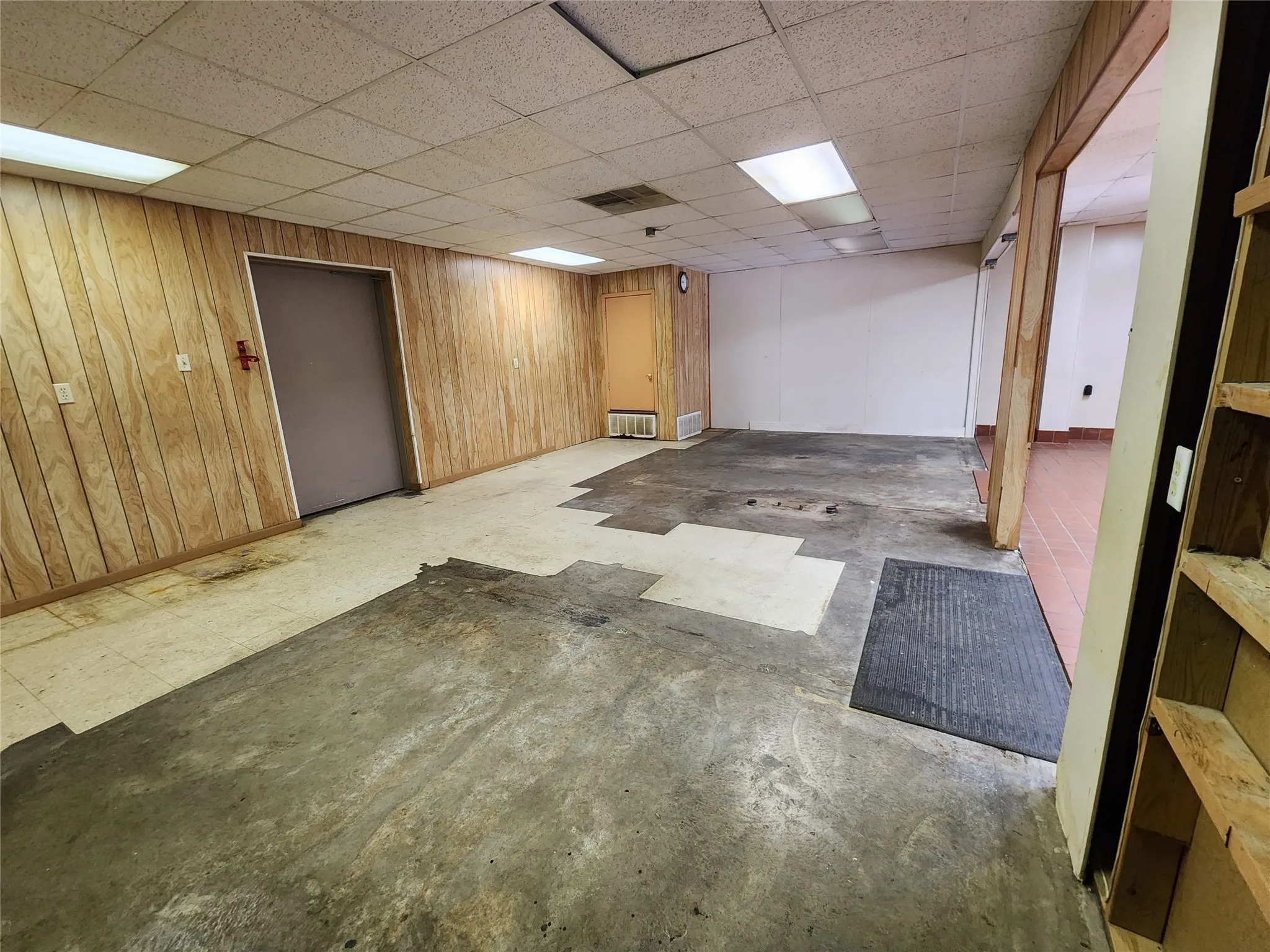 This space features wood paneling, an acoustic tile ceiling with fluorescent lighting, and a combination of concrete, tiled, and exposed flooring