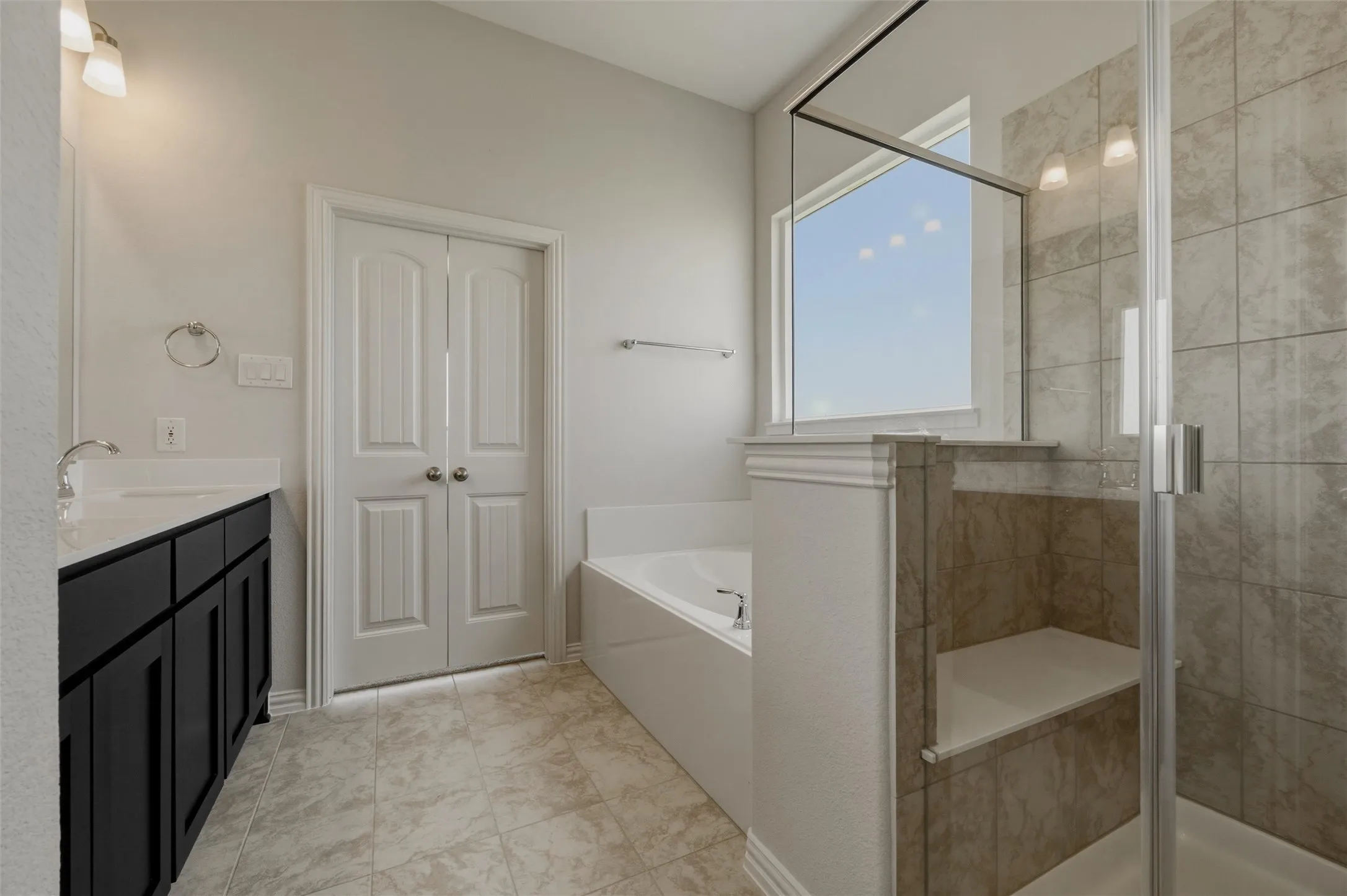 Spacious bathroom featuring a dual vanity with dark cabinetry, an enclosed shower with a built-in bench, a separate soaking tub, and tile flooring