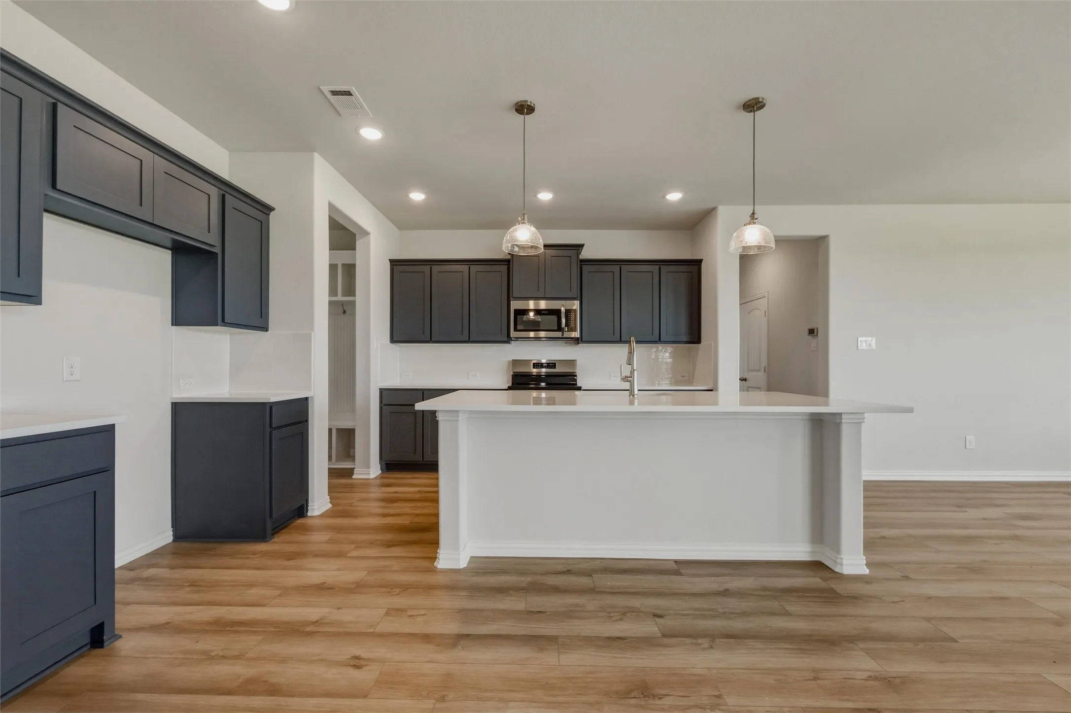 Kitchen featuring dark cabinetry, stainless steel appliances, a center island with a built-in sink, pendant lighting, and wood-finish flooring