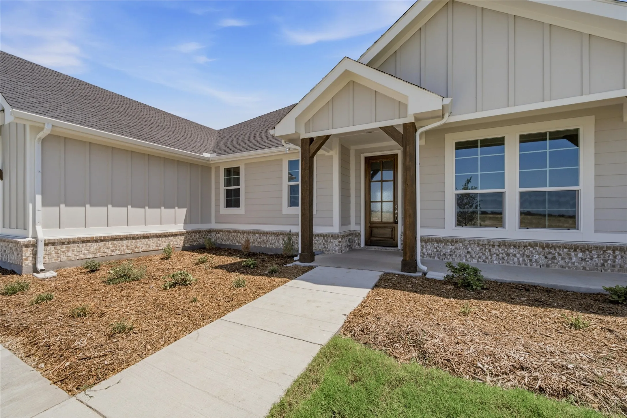 Stone-look exterior siding, wood-finish columns, dark wood entry door with glass panels, white-framed windows, and a concrete pathway