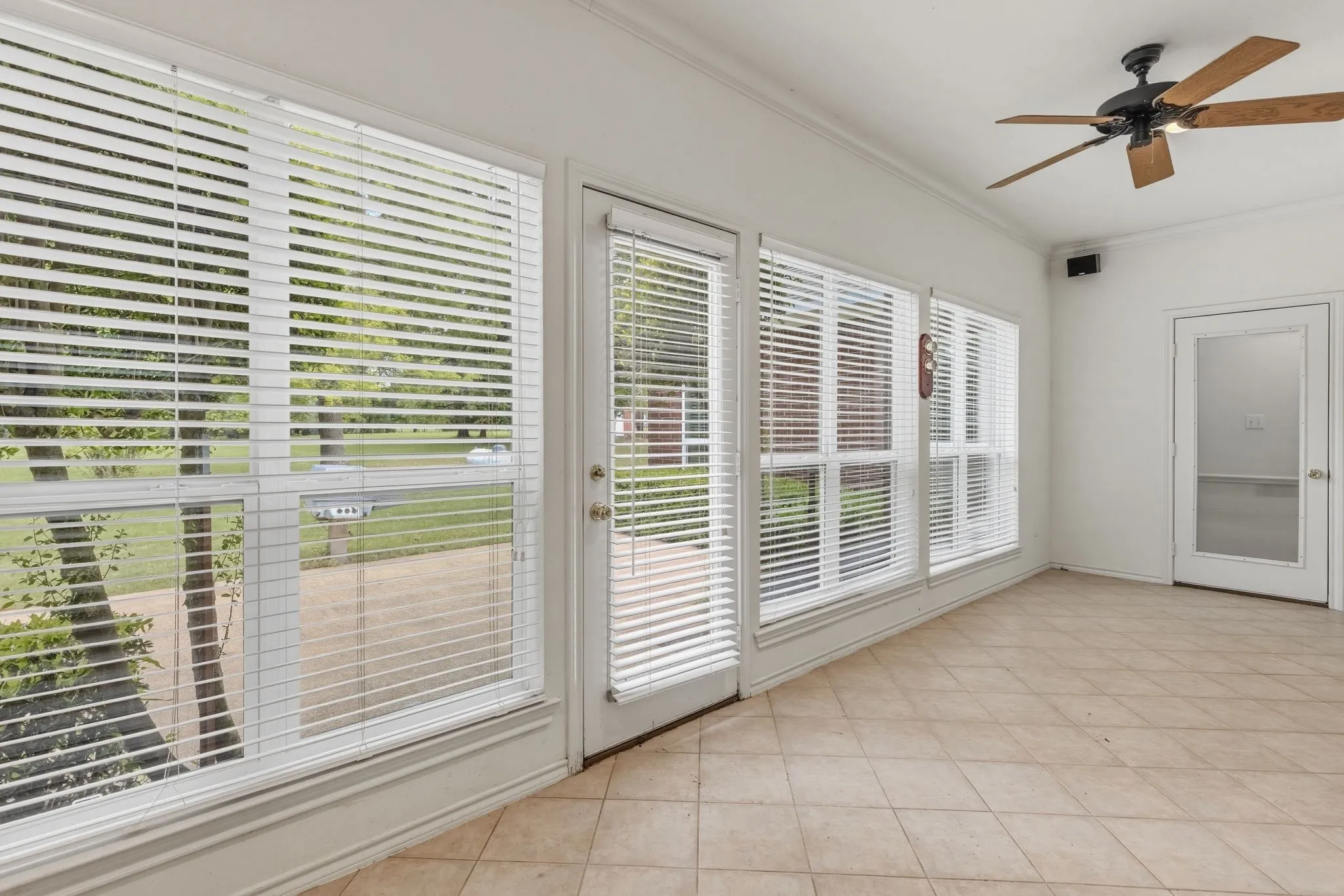 This room features large windows with white blinds, a tiled floor, and a ceiling fan with wooden blades