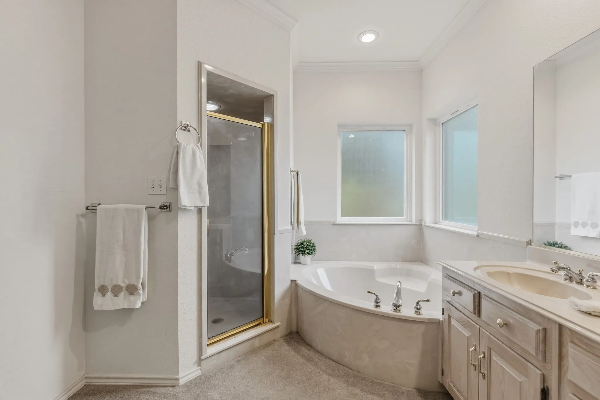 The bathroom features a corner soaking tub, a separate shower with a gold-toned frame, a vanity with an integrated sink, and windows providing natural light