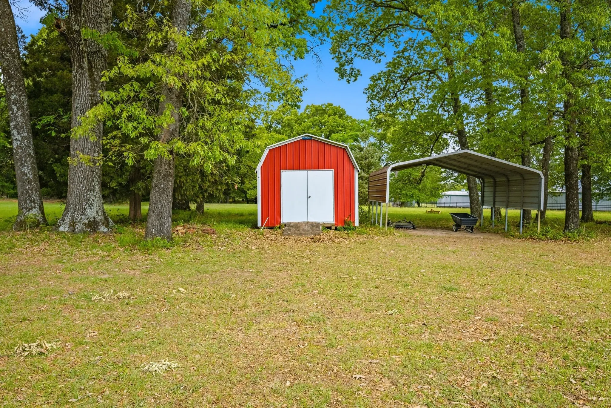 The property features a red storage shed with white doors and a metal carport, set within a spacious yard with mature trees