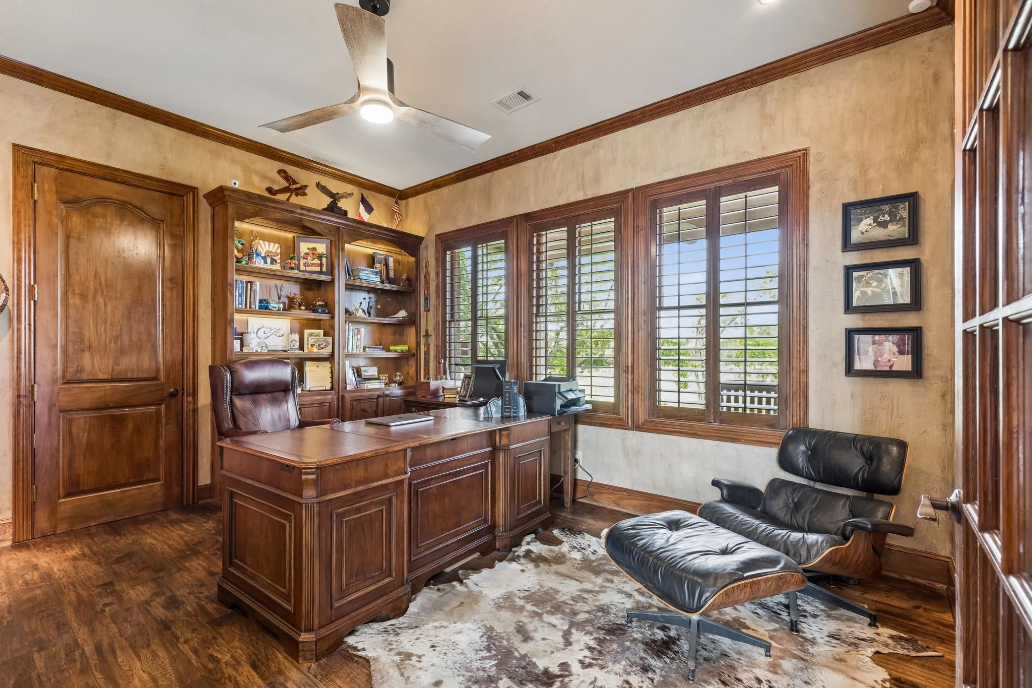 The room features wood flooring, a built-in wooden bookshelf with lighting, and three windows with wooden blinds