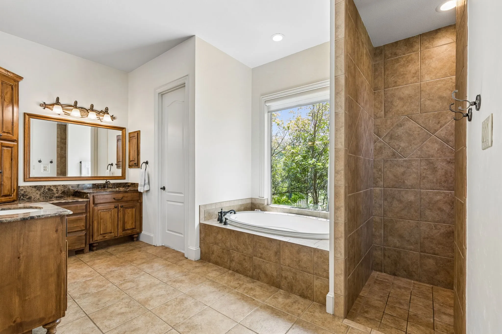 This bathroom features tile flooring, a built-in tub with a window view, a tiled shower, and a double vanity with wooden cabinetry and stone countertops
