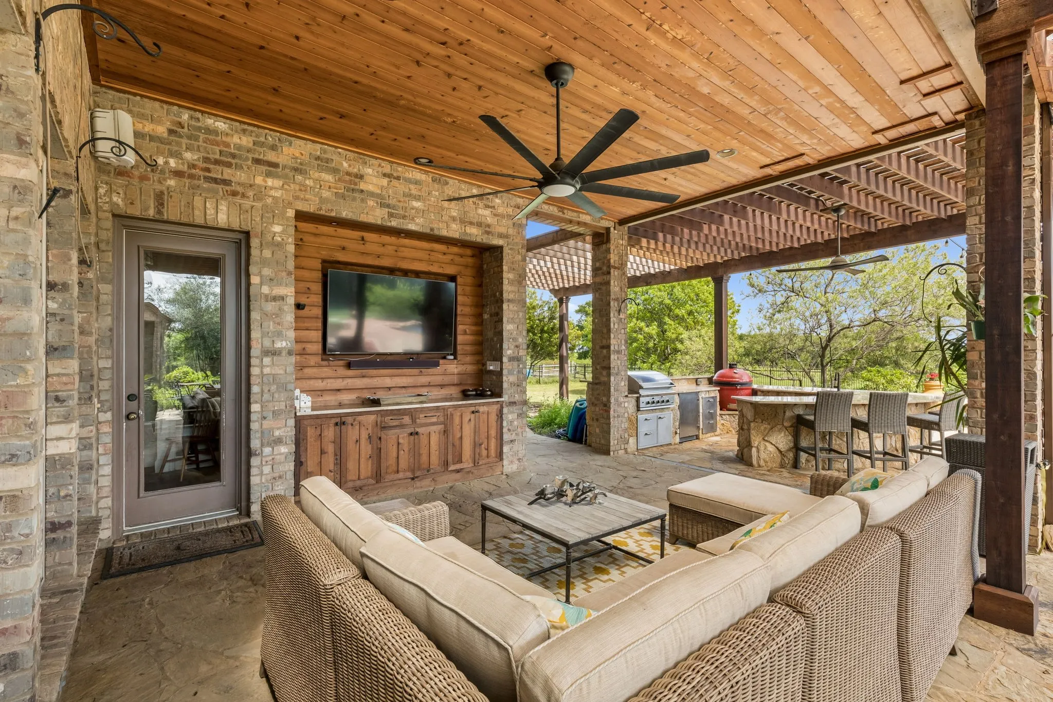Expansive covered patio featuring a wood-paneled ceiling with ceiling fans, a built-in media center with storage, and an outdoor kitchen with a stone-clad bar area