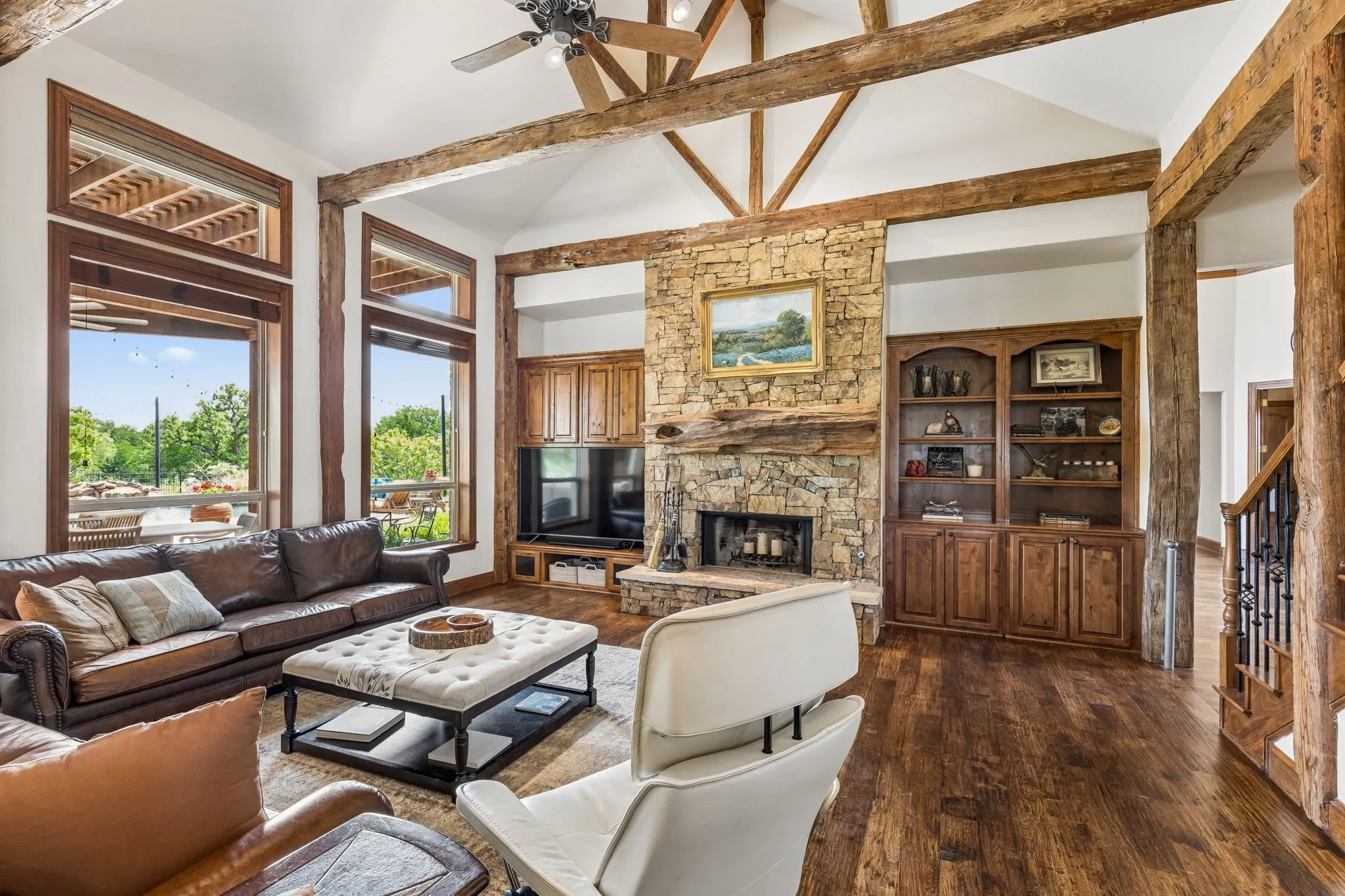 This expansive living area features vaulted ceilings with exposed wood beams, large windows providing abundant natural light and views of greenery, and rich hardwood flooring
