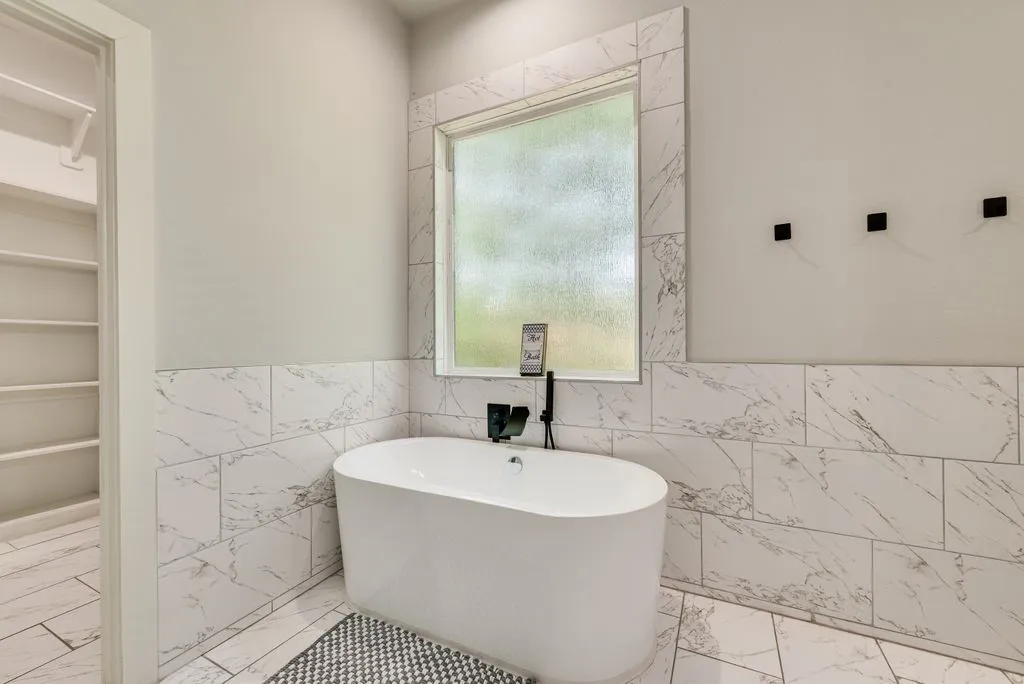 The bathroom features a modern freestanding bathtub, white marble-patterned wall and floor tiling, and a window providing natural light