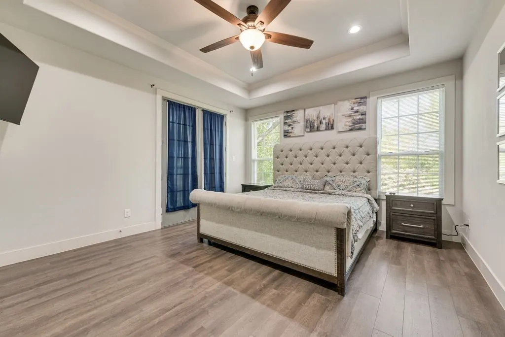 This bedroom features wood-look flooring, a tray ceiling with a ceiling fan, and multiple windows providing natural light