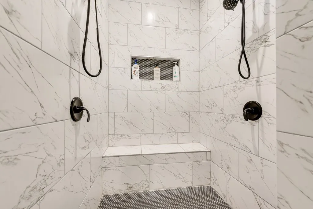 Spacious shower featuring white marble-look tile walls, dual shower heads with matte black fixtures, a built-in bench, and a recessed niche for storage
