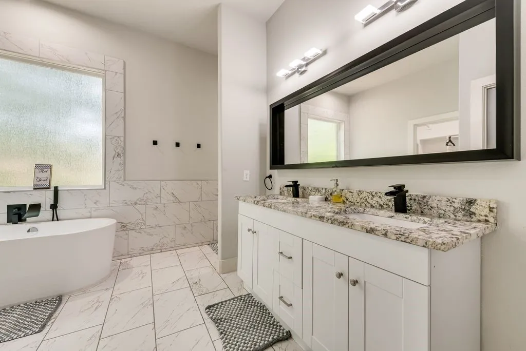 Spacious bathroom featuring a double vanity with granite countertops and a large mirror, a standalone bathtub with a dark faucet, and tiled floors