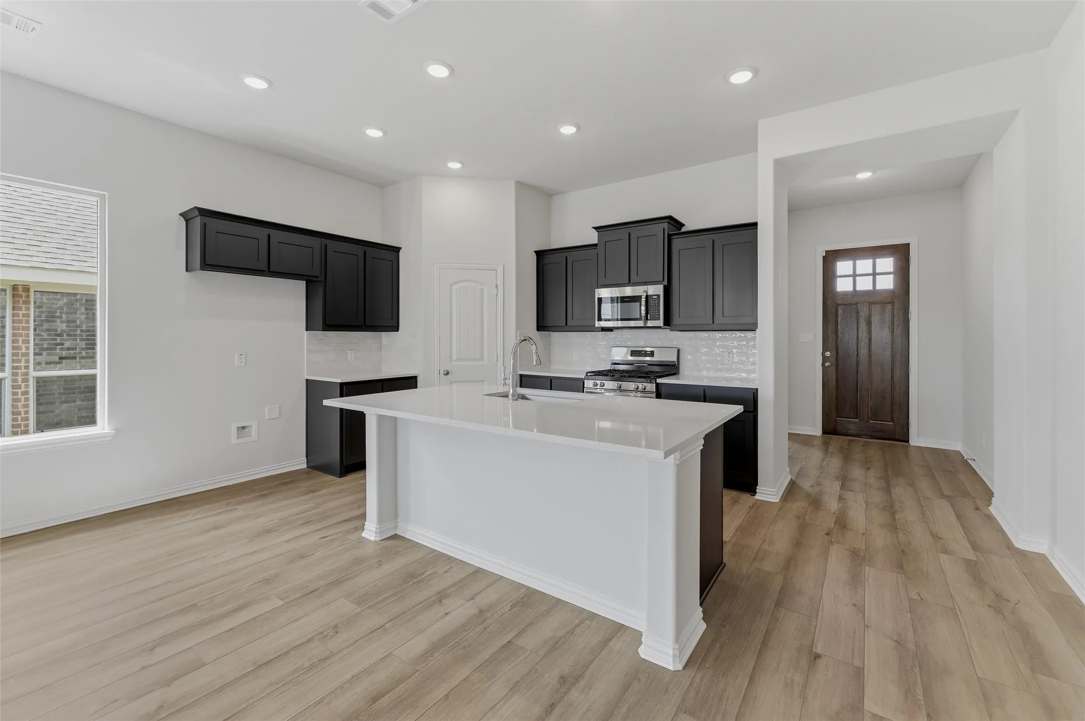 Open-concept kitchen featuring an island with a white countertop, dark cabinetry, stainless steel appliances, and wood-finish flooring