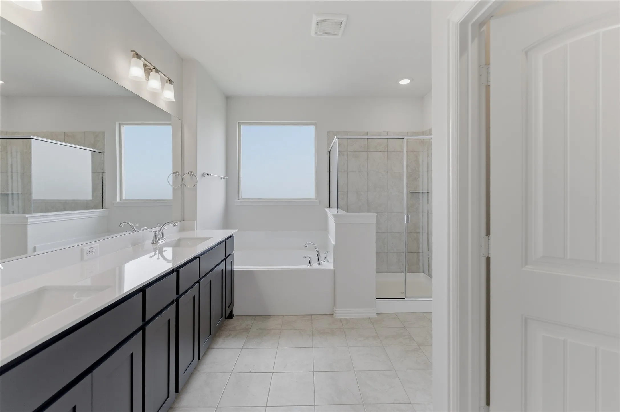 Contemporary bathroom featuring a long double vanity with dark cabinetry and light countertops, a built-in soaking tub, a separate glass-enclosed shower with tiled walls, and light tile flooring
