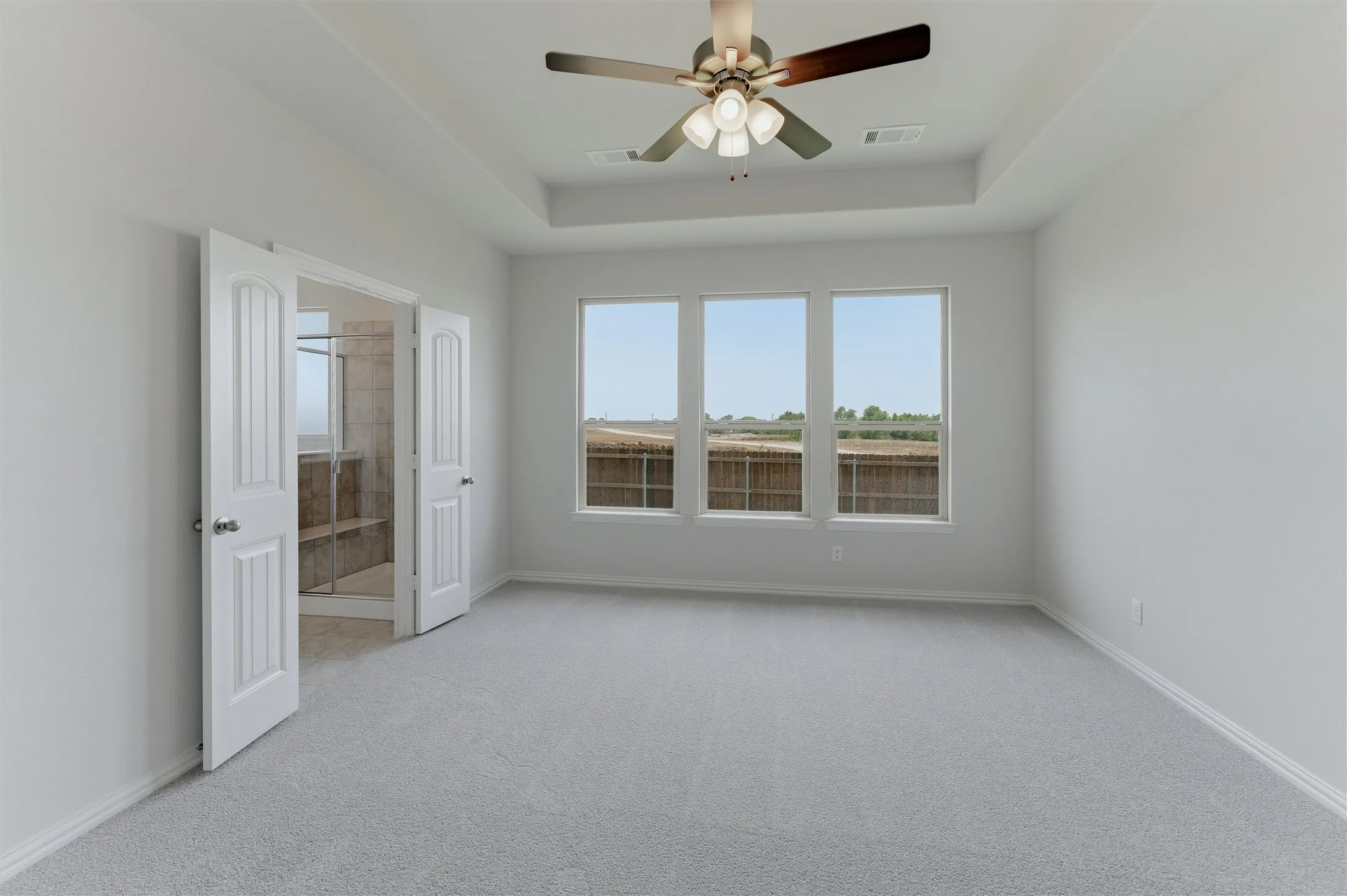 Tray ceiling with a ceiling fan, three windows overlooking a privacy fence, light gray carpet, and white trim