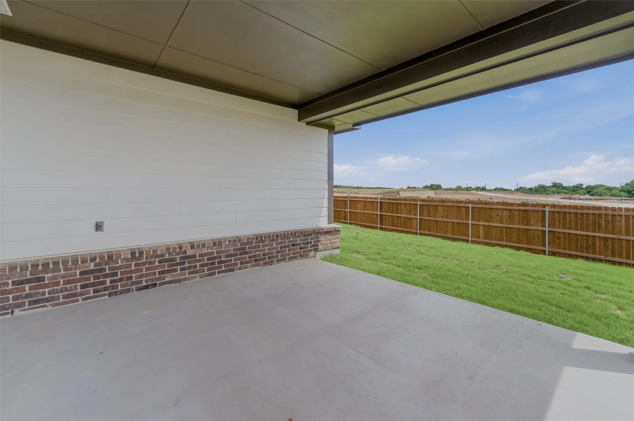 Covered concrete patio with white siding and brick veneer, bordered by a wooden privacy fence and a grass lawn