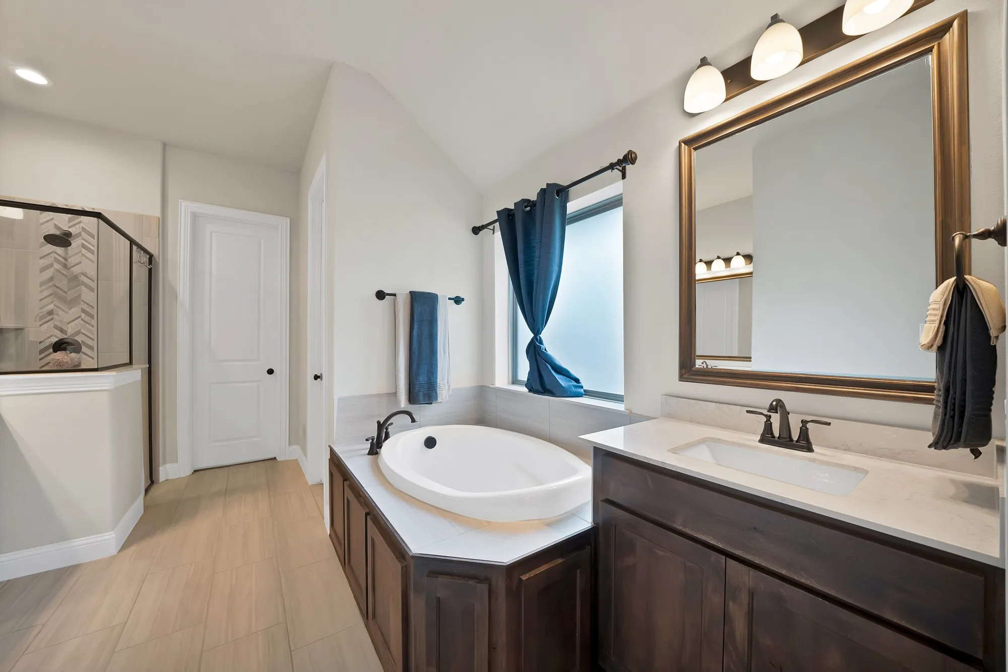 The bathroom features a built-in tub, a dark wood vanity with a white countertop and rectangular sink, and a glass-enclosed shower with chevron-patterned tiling