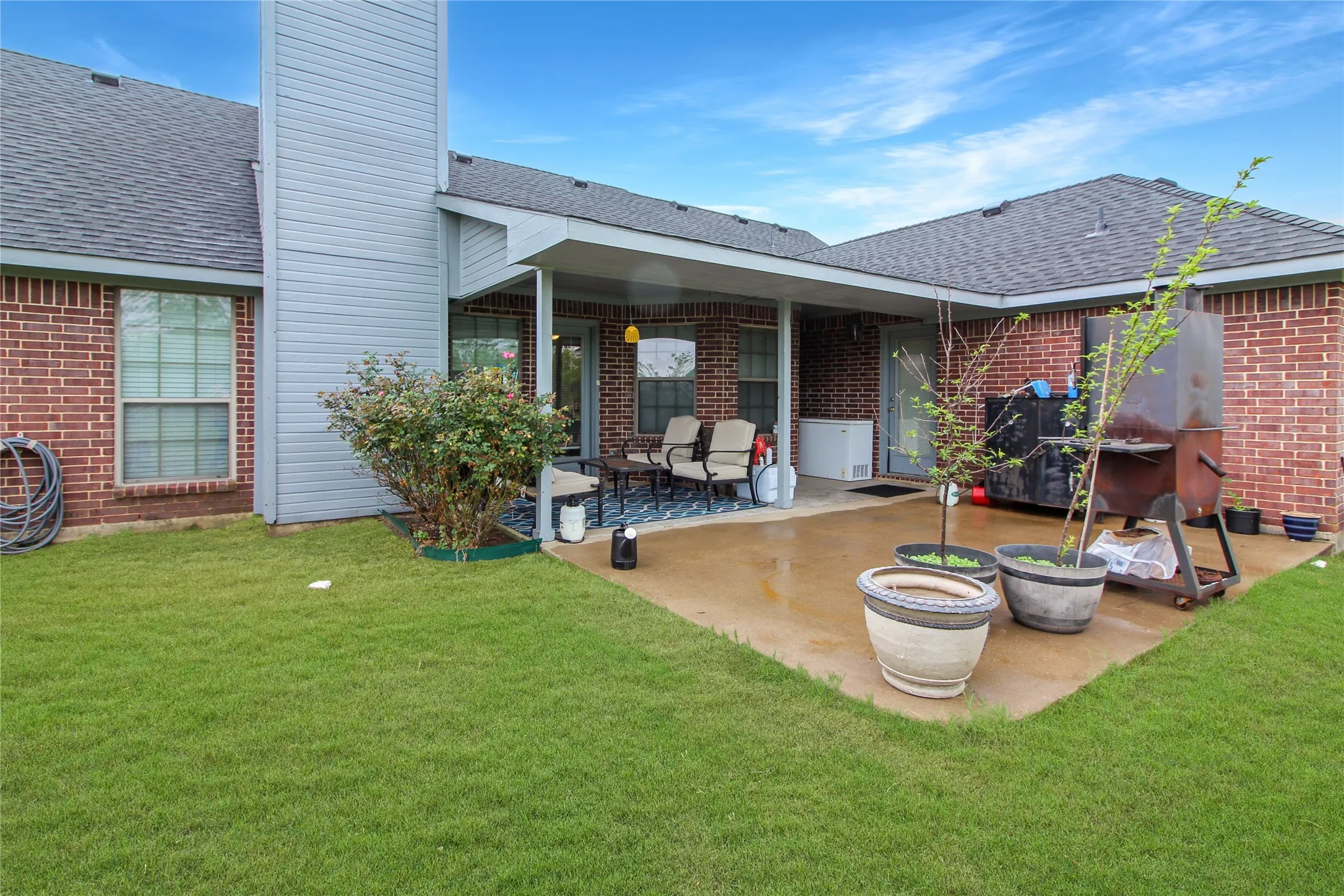 Expansive grassy backyard featuring a covered patio, red brick exterior, dark shingle roof, and a prominent chimney structure