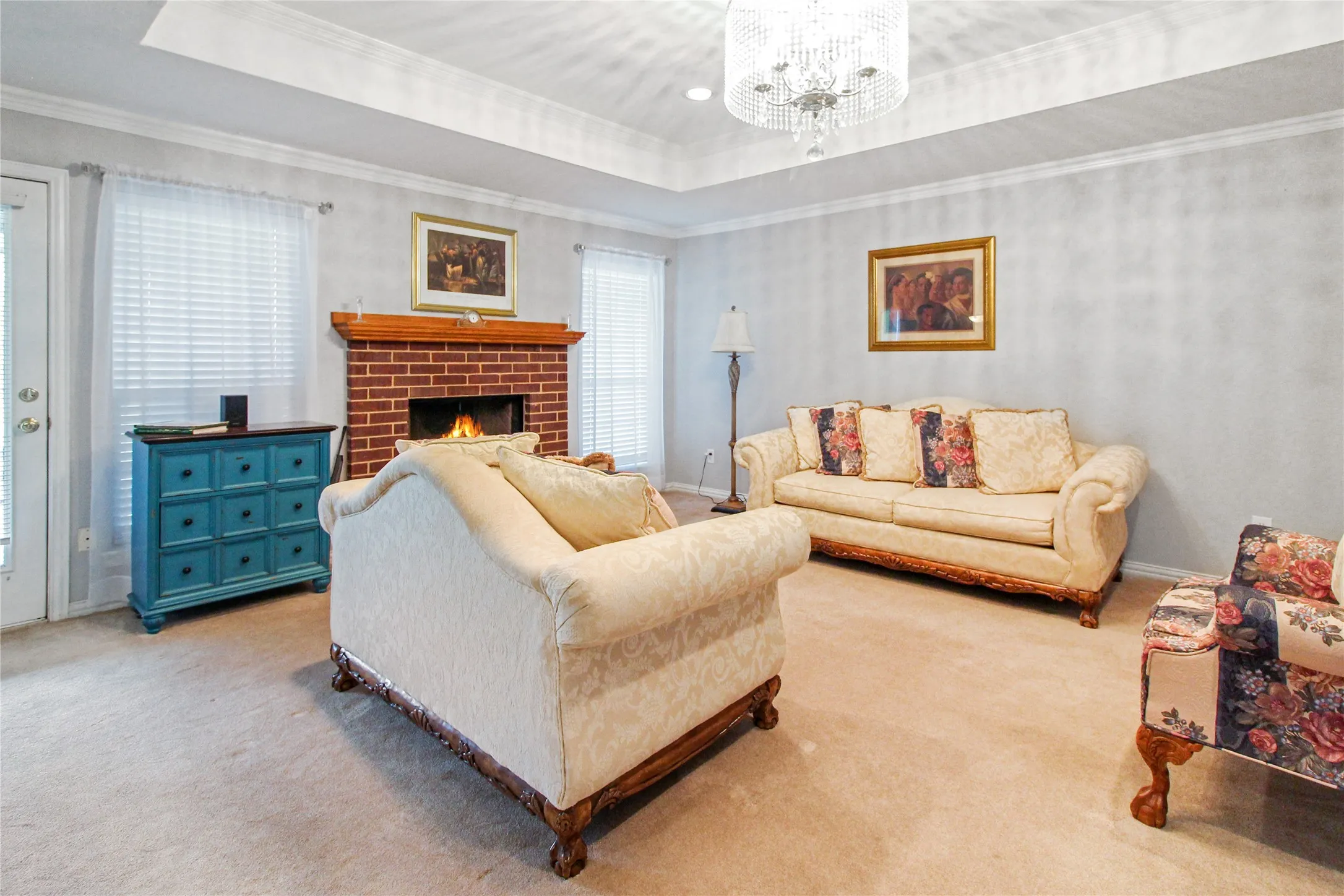 Living area featuring a brick fireplace with wood mantel, recessed tray ceiling, crown molding, and a chandelier lighting fixture